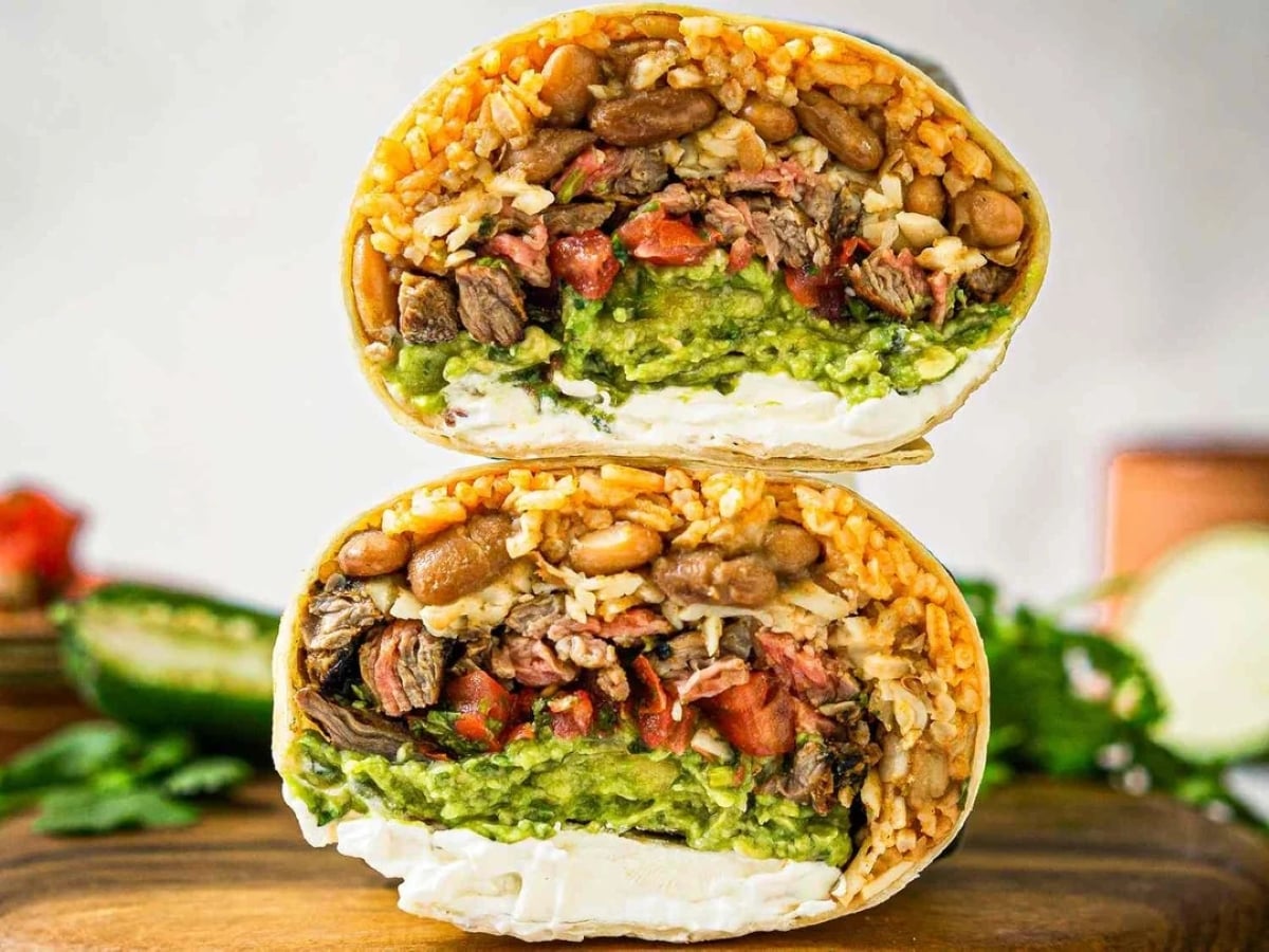 A close-up of a burrito cut in half, showing layers of rice, beans, steak, guacamole, tomatoes, and sour cream, stacked on a wooden surface with blurred ingredients in the background.