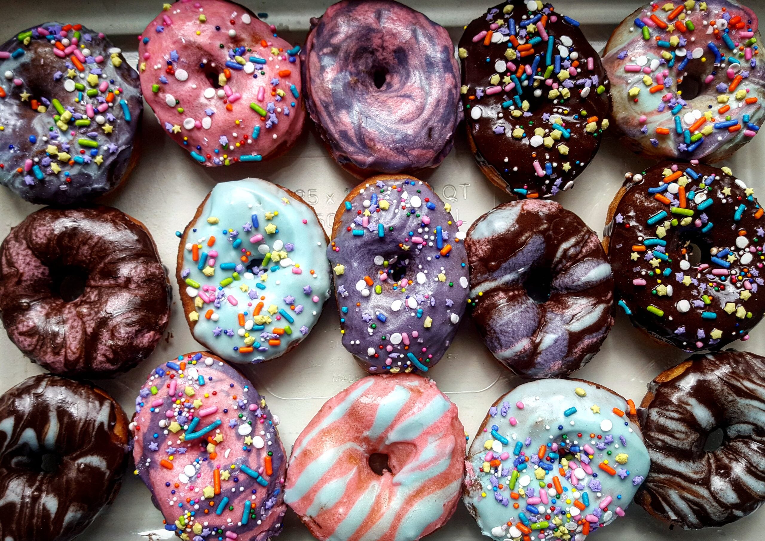 A box of fifteen colorful donuts with various pastel icing and sprinkles, arranged in three rows. The icing colors include pink, blue, purple, and chocolate, with swirl and sprinkled designs.