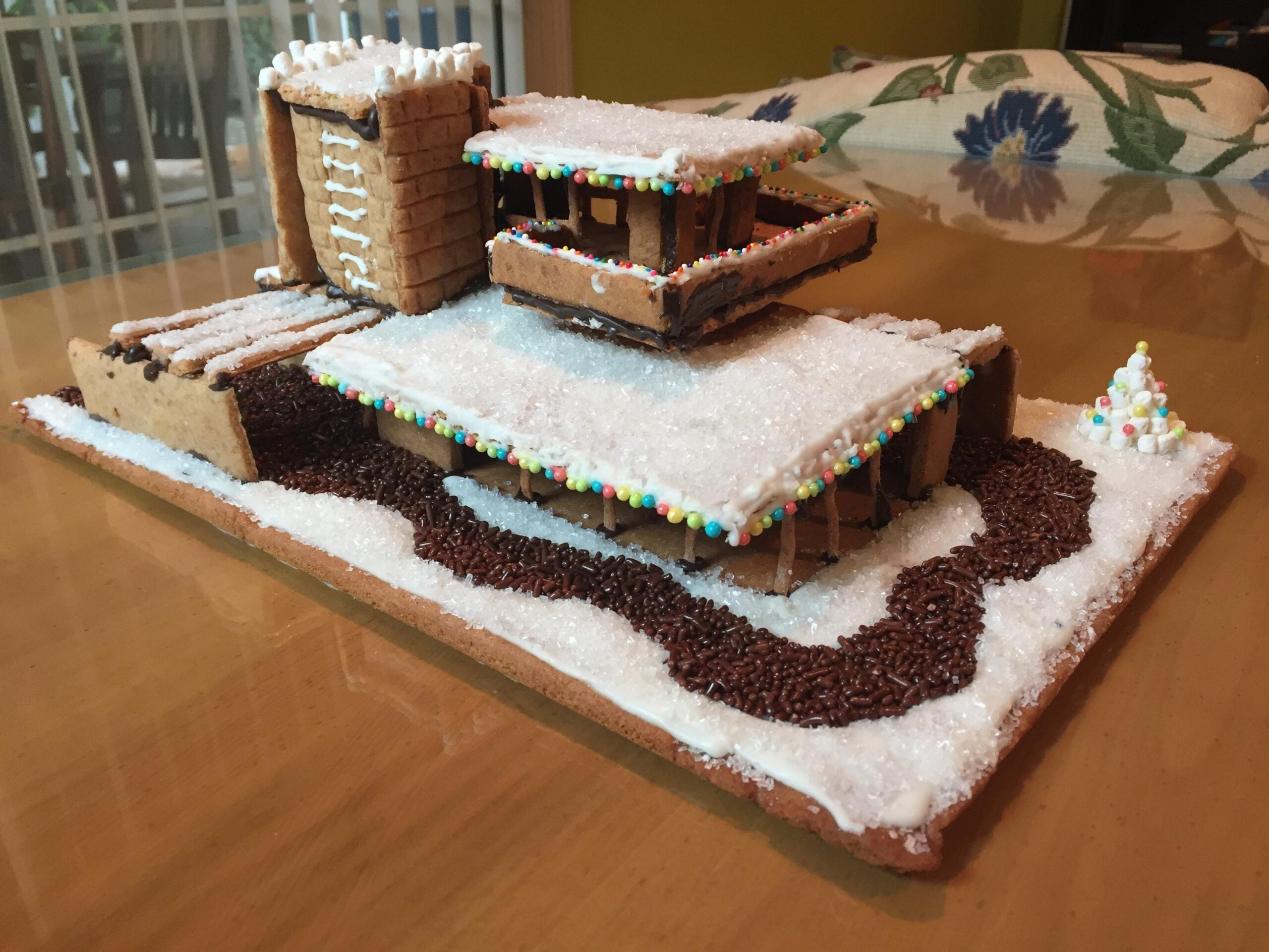 A gingerbread house modeled after a modern building, decorated with white icing, candy, and chocolate sprinkles, sits on a brown surface with a snowy effect and a small candy tree—an inviting display of homemade Christmas treats.