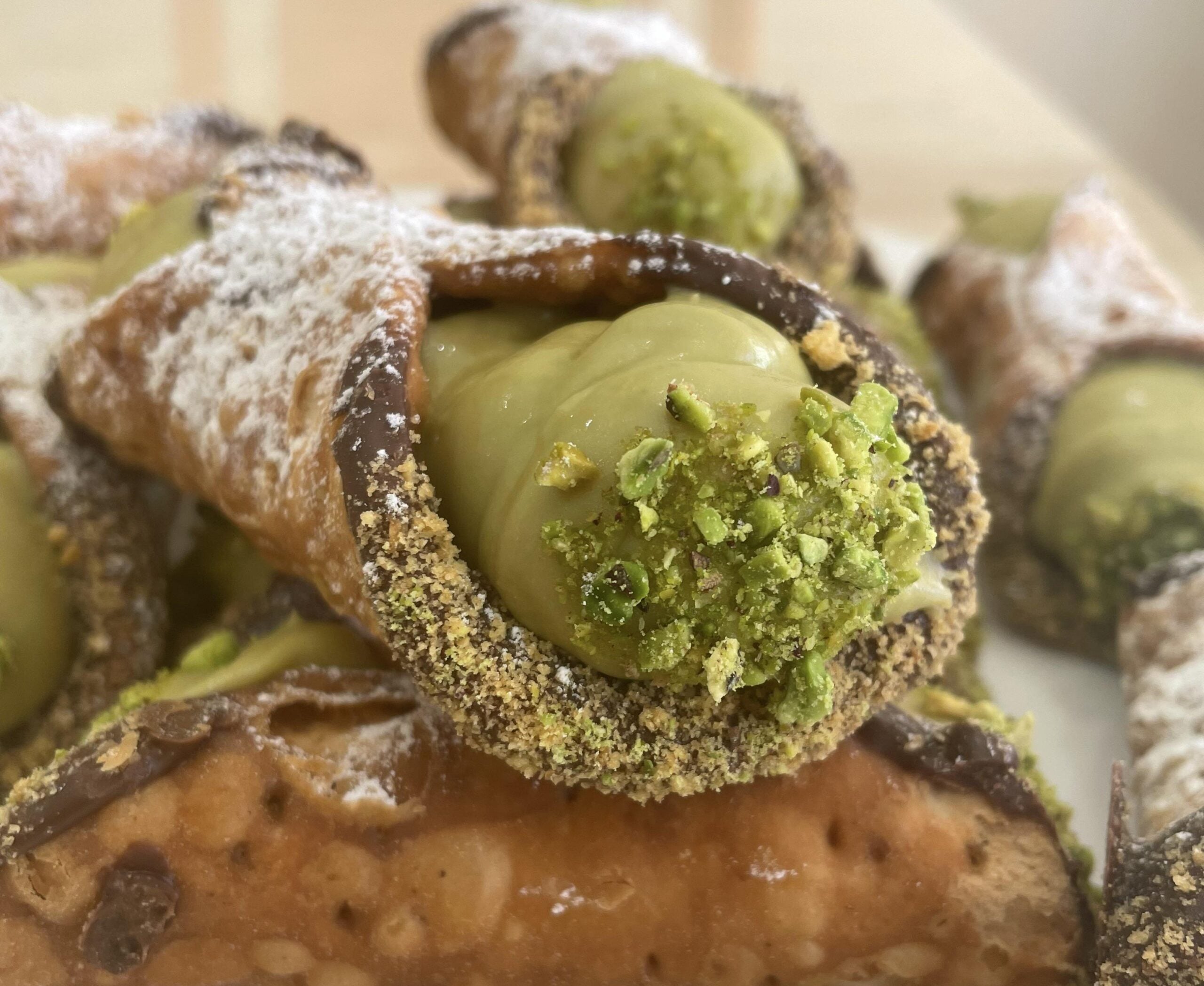 Close-up of homemade cannolis filled with pistachio cream, dipped in chocolate and chopped pistachios, and dusted with powdered sugar. Other cannoli are blurred in the background.
