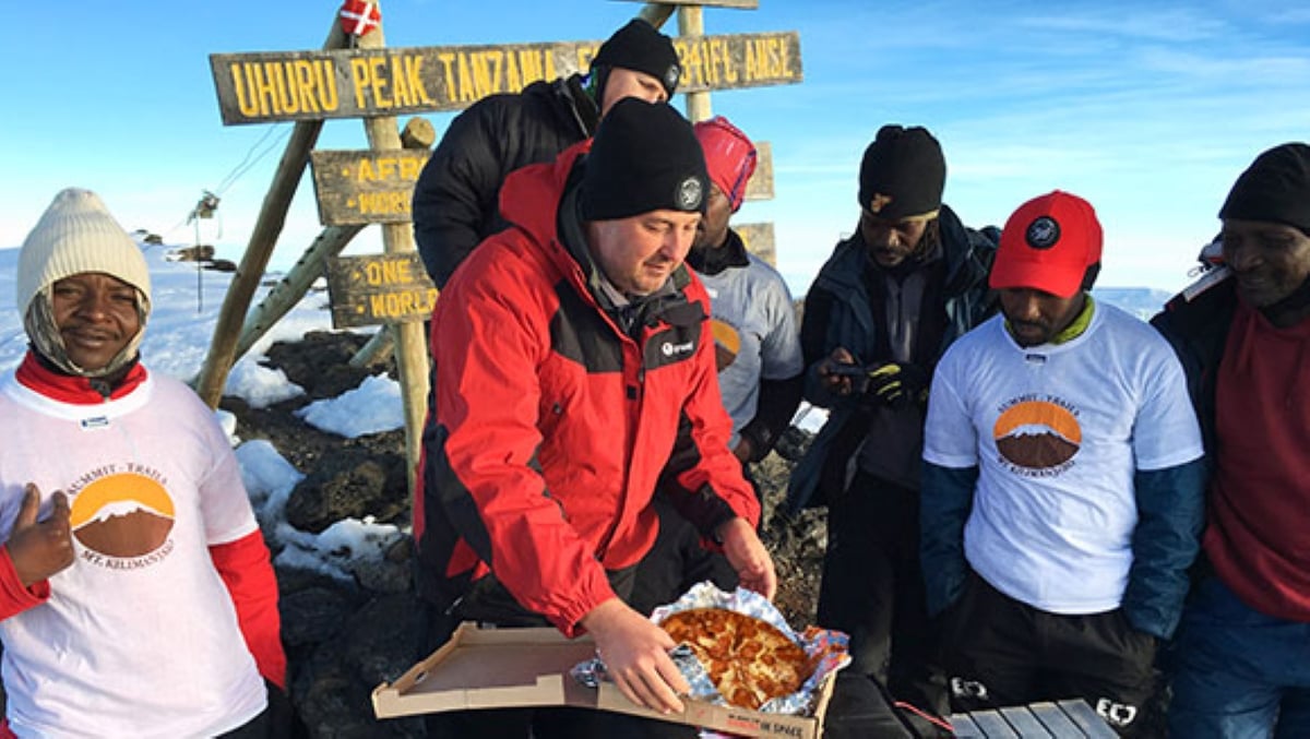 A group of climbers at Uhuru Peak on Mount Kilimanjaro gather around a man holding a pizza box, with summit signs and snow in the background—a truly unforgettable meal that makes for one of those Wild Fast-Food Facts you’ll want to share.