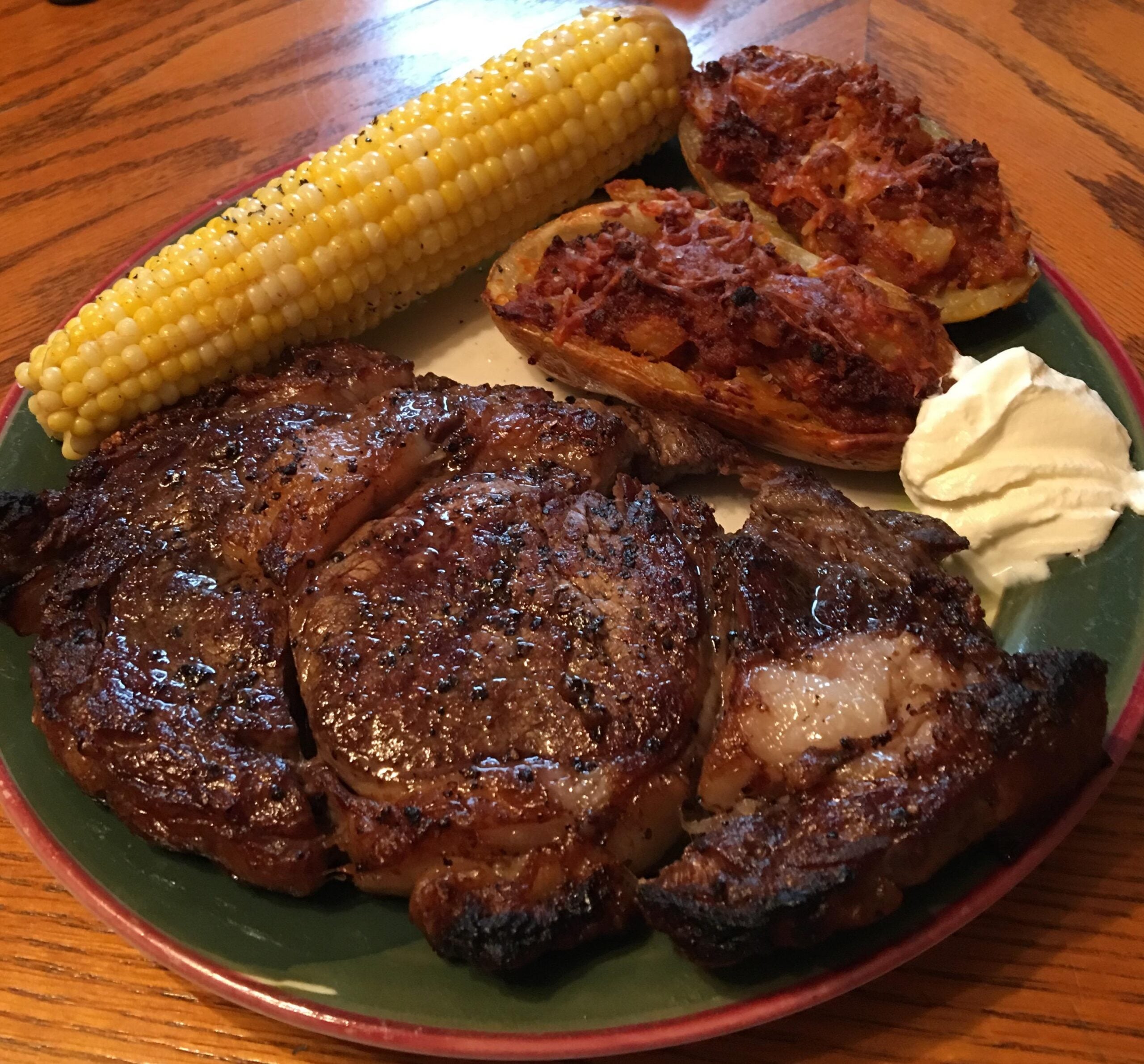A plate with grilled steak, a corn on the cob, two stuffed potato halves with bacon bits, and a scoop of sour cream on the side, set on a wooden table.