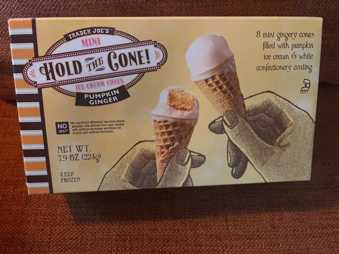 Box of Trader Joe’s “Hold The Cone!” mini pumpkin ginger ice cream cones, showing two hands holding cones with creamy filling. Text highlights pumpkin ice cream and ginger cones with white coating.