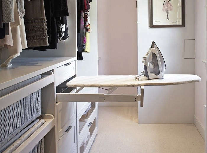 A pull-out ironing board with an iron is extended from a white built-in closet unit in a well-organized laundry or dressing room. Clothes hang on one side and a framed artwork hangs on the wall.