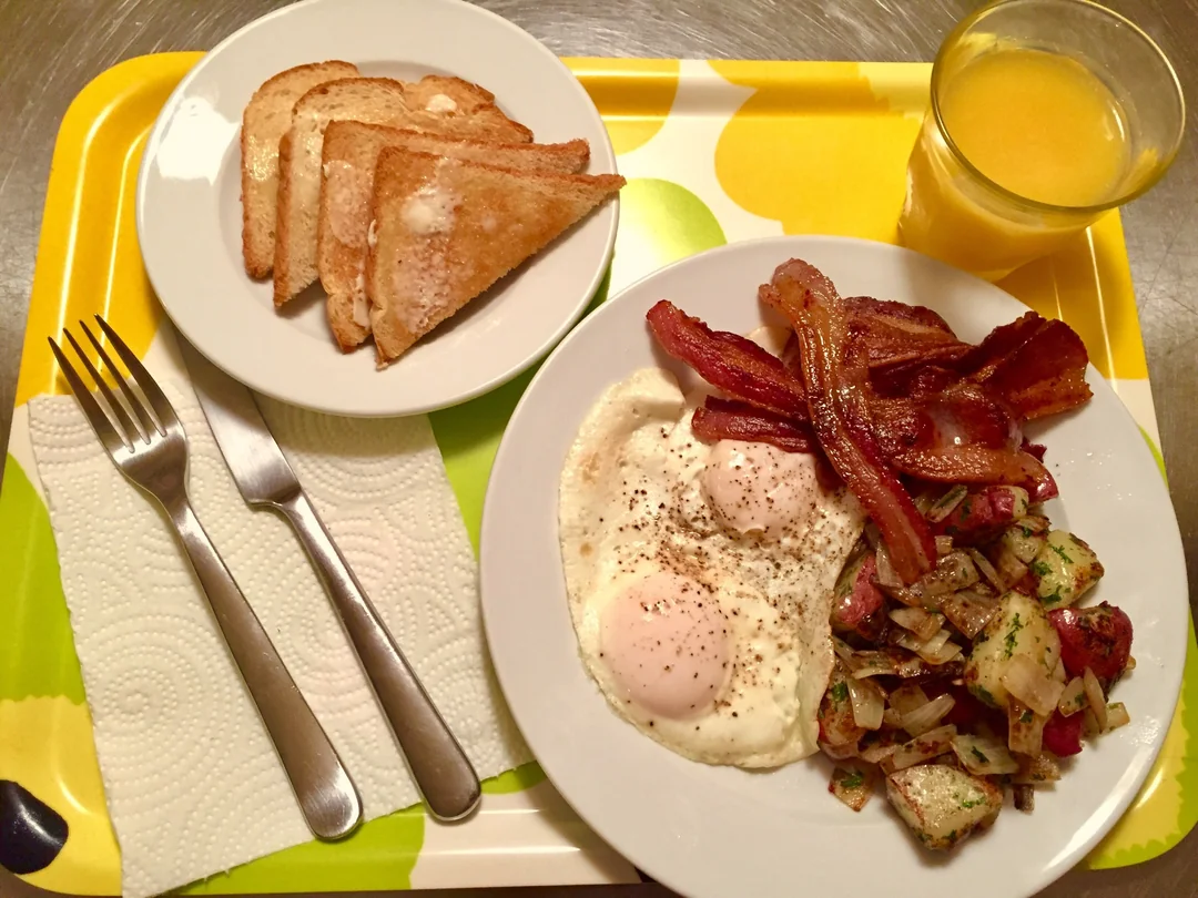 Breakfast tray with toast, fried eggs, bacon, seasoned potatoes, and a glass of orange juice. Fork and knife rest on a napkin beside the plate.