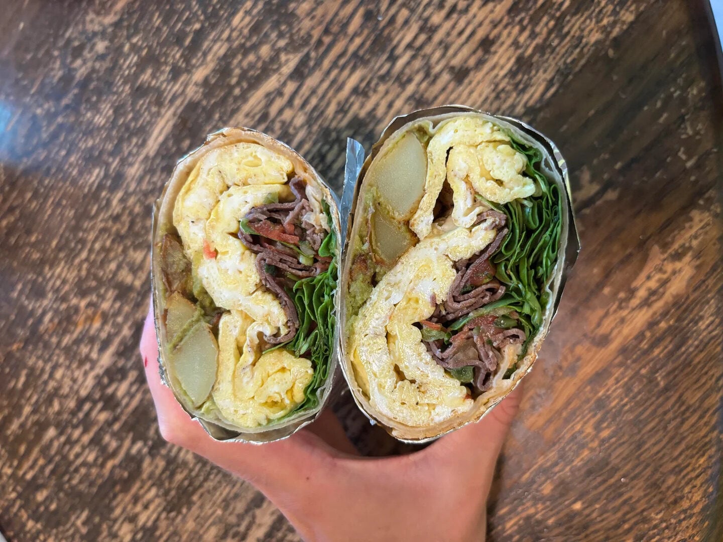 Two hands hold a halved breakfast burrito filled with scrambled eggs, shredded beef, spinach, and potato slices, wrapped in foil and placed on a wooden table.