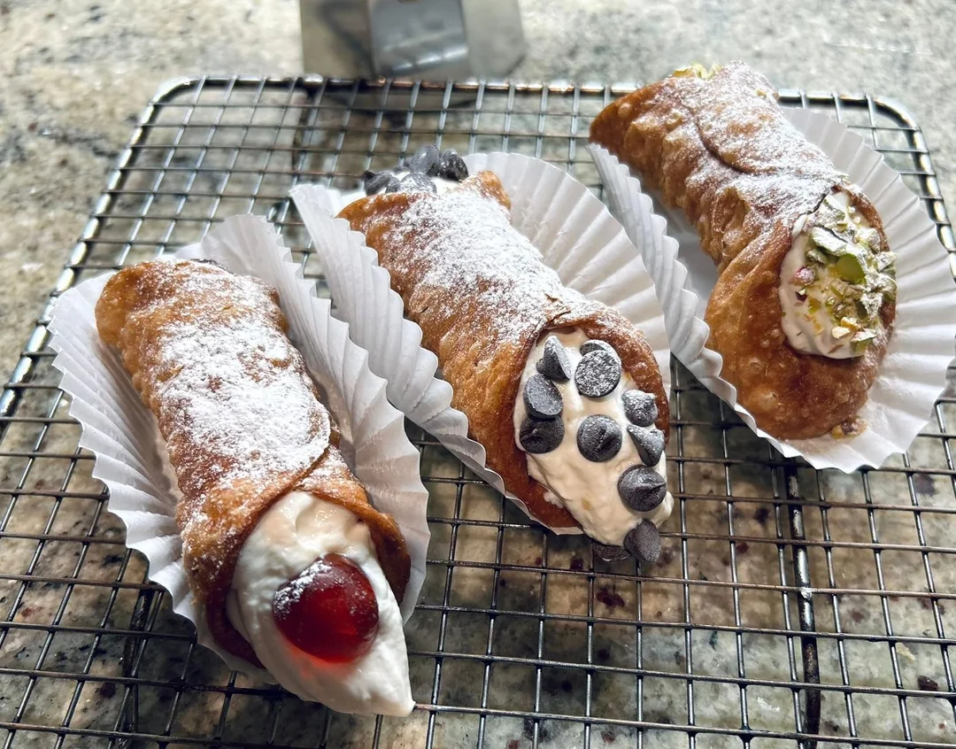 Three homemade cannolis rest on a wire rack, each dusted with powdered sugar. Fillings include a red cherry, chocolate chips, and chopped pistachios. Each cannoli sits in a white paper tray.