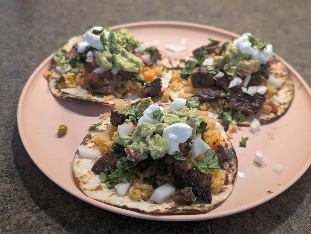 Three tacos on a pink plate are topped with grilled meat, rice, onions, cilantro, guacamole, and sour cream, set on a brown countertop.