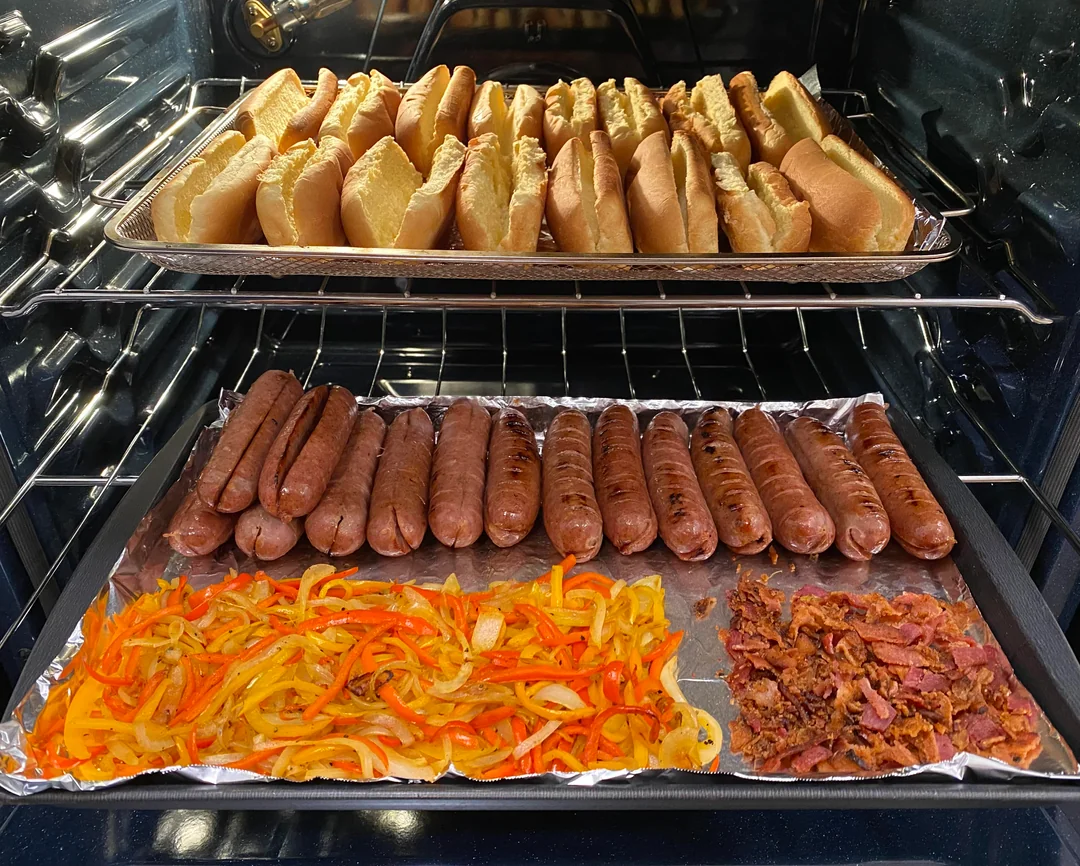 Hot dog buns on a baking tray in the top oven rack, with sausages, grilled onions and peppers, and crumbled bacon on foil-lined baking sheet on the lower oven rack.