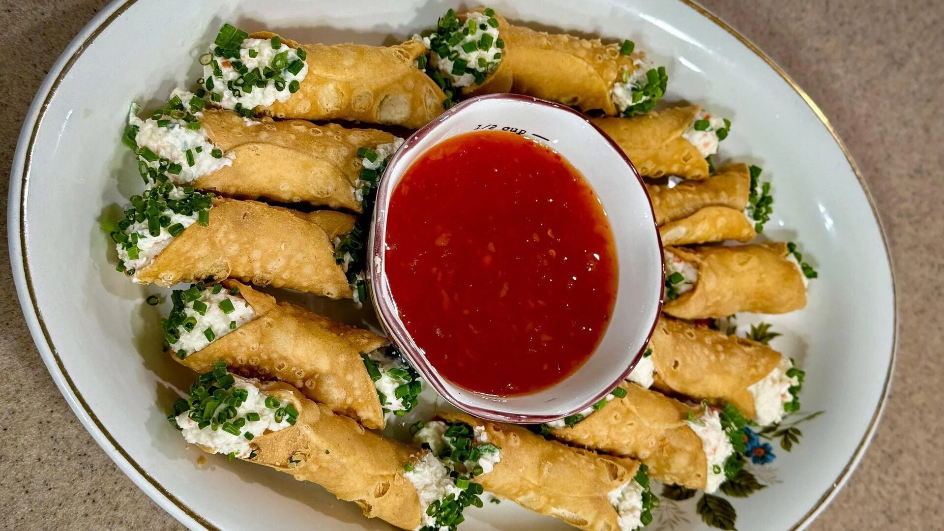 A plate of crispy homemade cannoli shells filled with a creamy mixture, garnished with chopped herbs, arranged in a circle around a bowl of red dipping sauce.