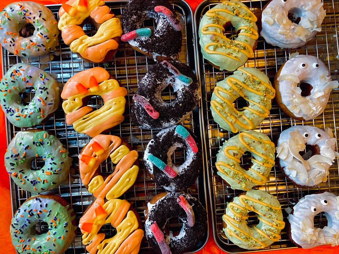 Twelve decorated donuts on cooling racks, topped with colorful icing, sprinkles, gummy worms, and candy eyes, creating a playful and festive appearance.