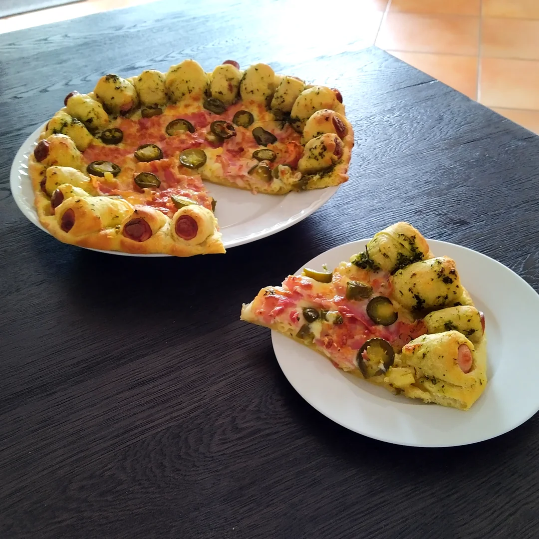 A pizza with a crust made of hot dog pieces, topped with ham, green olives, and herbs, sits on a plate. One slice is served on a separate white plate on a dark table.