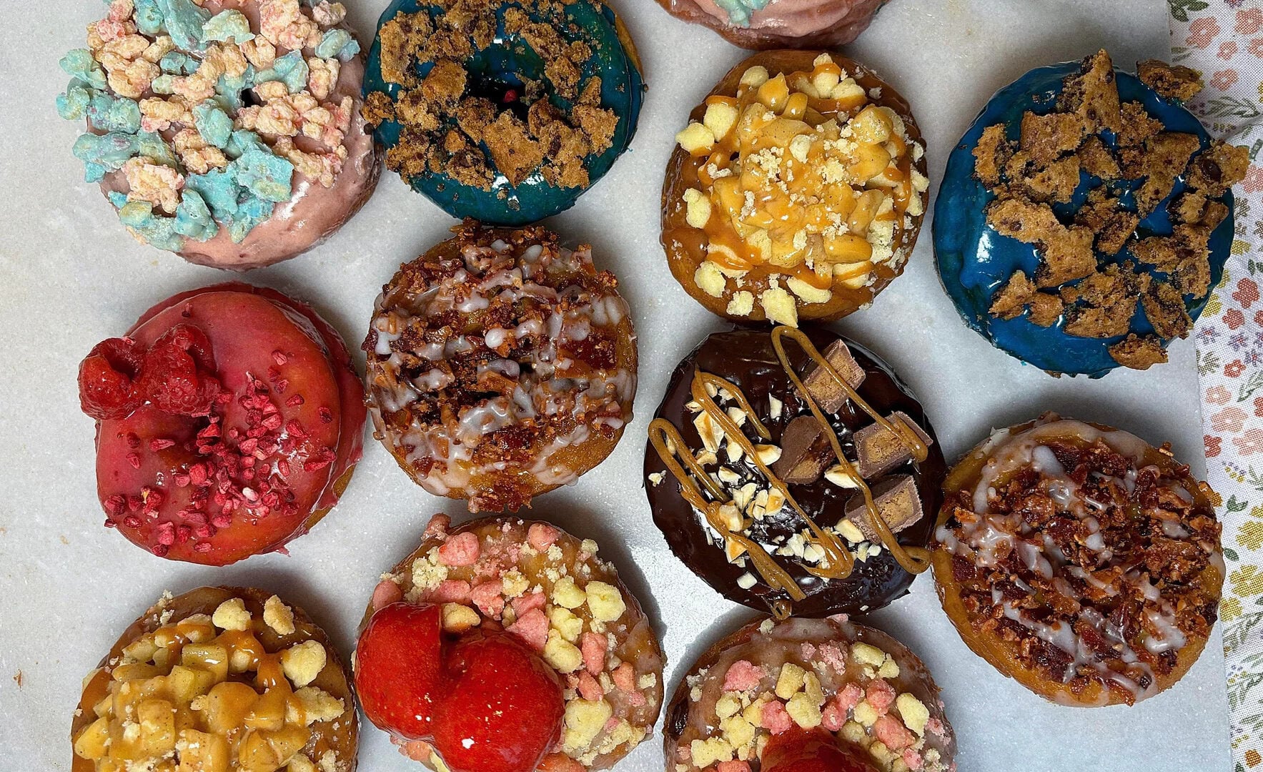 A selection of twelve colorful donuts, each topped with various toppings like fruit, candy, crumbles, and drizzle, arranged in rows on a white surface.
