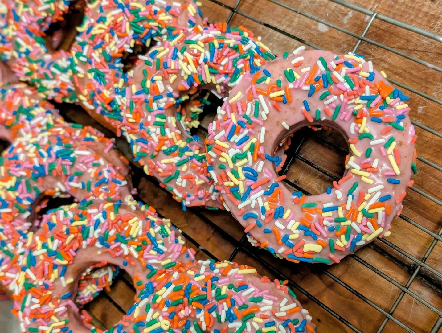 Rows of donuts with pink icing and colorful rainbow sprinkles are cooling on a wire rack over a wooden surface.