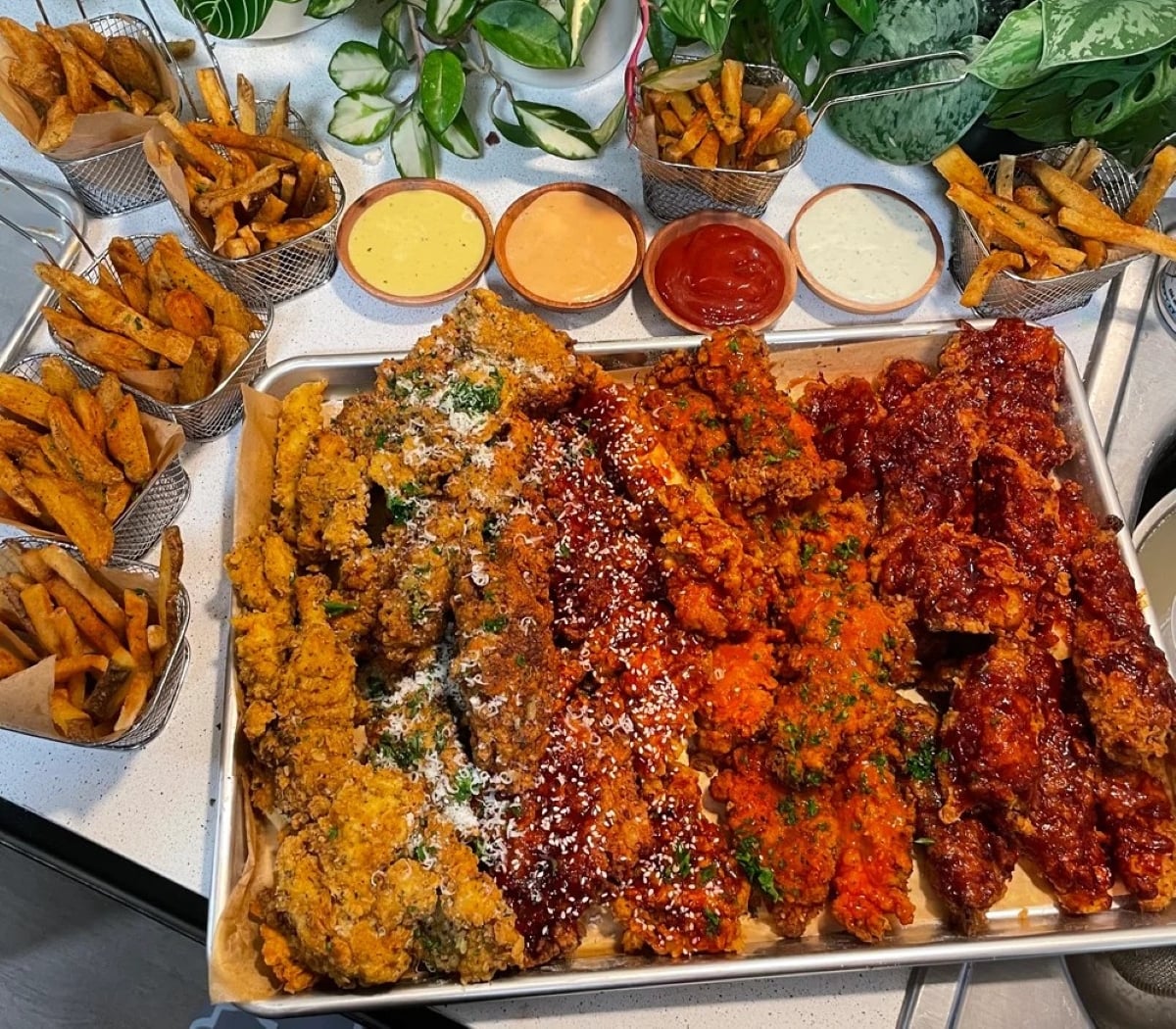 A tray of assorted fried chicken strips with various sauces is surrounded by baskets of seasoned fries. The scene is garnished with fresh herbs and leafy plants in the background.
