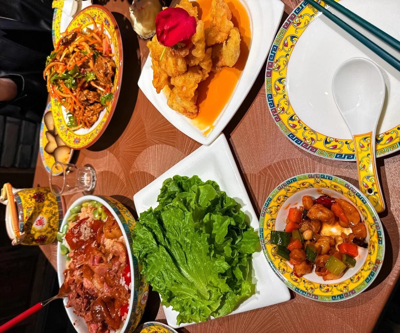 Colorful Chinese dishes on a table, including stir-fried vegetables, fried food with a red garnish, leafy lettuce, meat over rice, and a yellow ornate bowl, with chopsticks and plates—showcasing the amazing food people ate while on vacation.