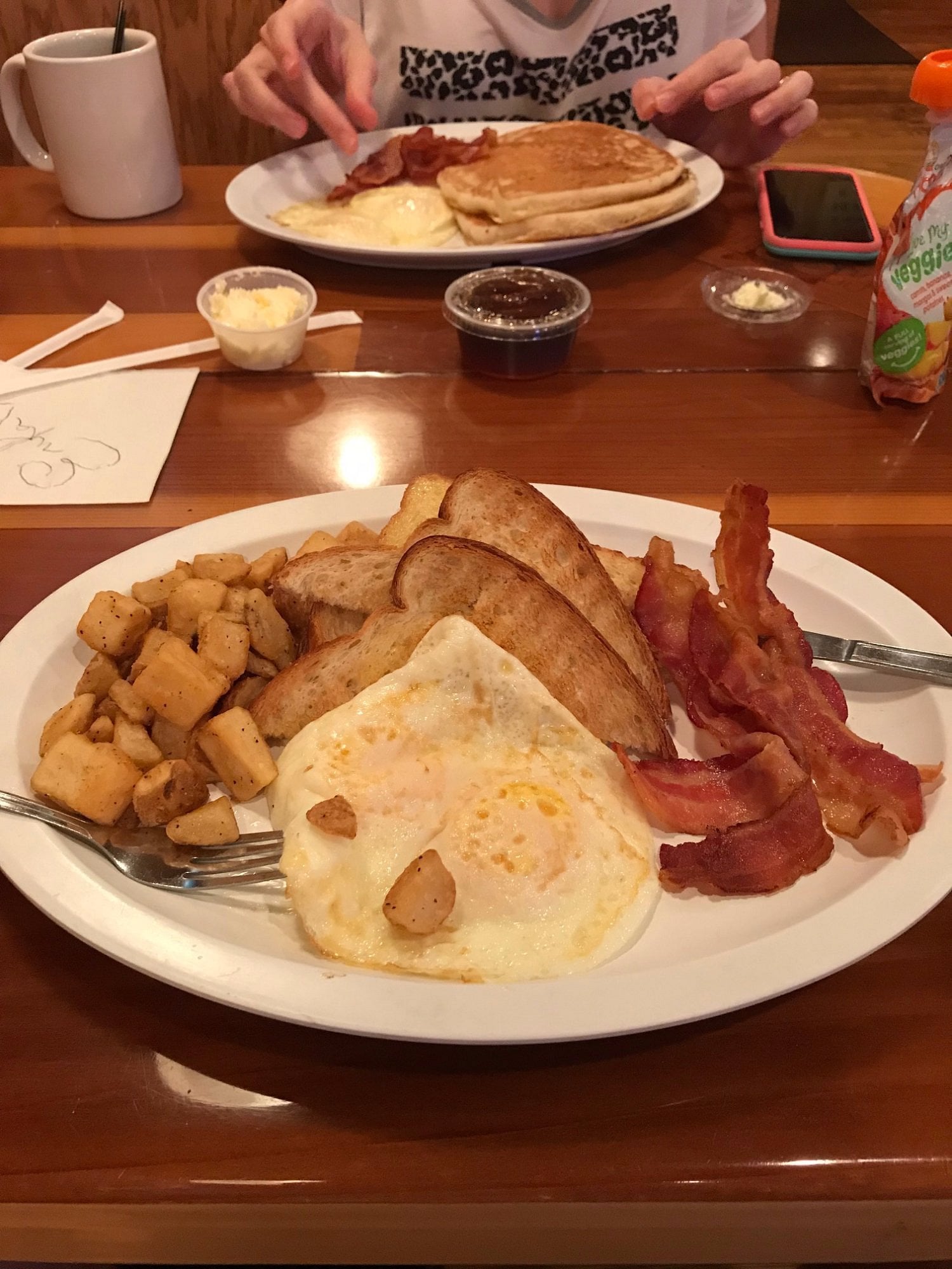 A plate of sunny-side-up eggs, bacon, toast, and home fries on a wooden table, with another plate of pancakes and a person’s hands in the background. Coffee, butter, and jam are also visible.