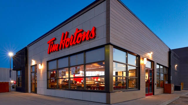 A Tim Hortons restaurant with large windows and red signage is pictured at dusk, with lights on inside and a mostly empty parking lot outside.