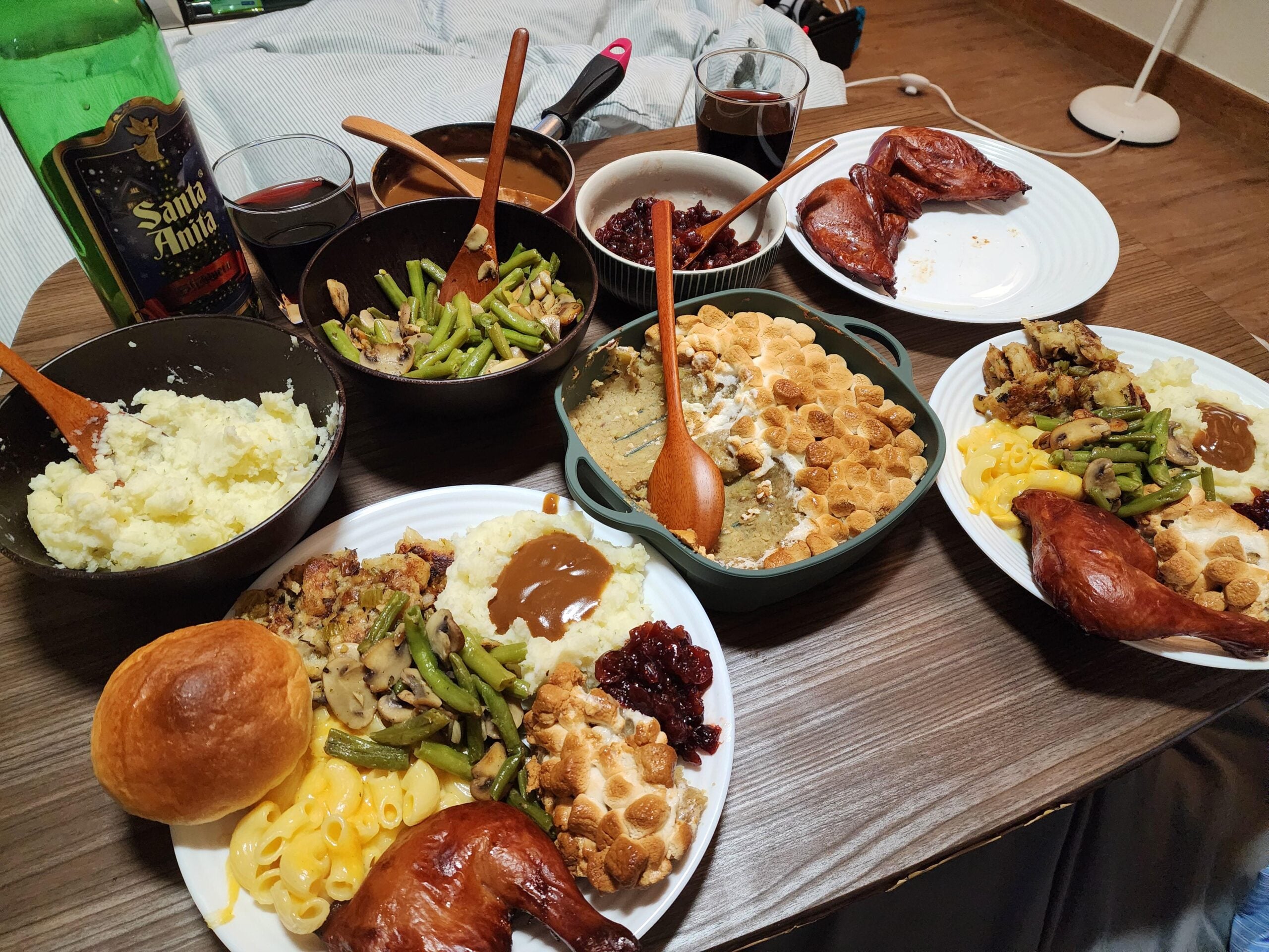 Two plates with roasted chicken, mashed potatoes, green beans, macaroni, bread, cranberry sauce, and sweet potato casserole. Two glasses of red drink, a beer bottle, bowls of sides, and utensils are also on the table.