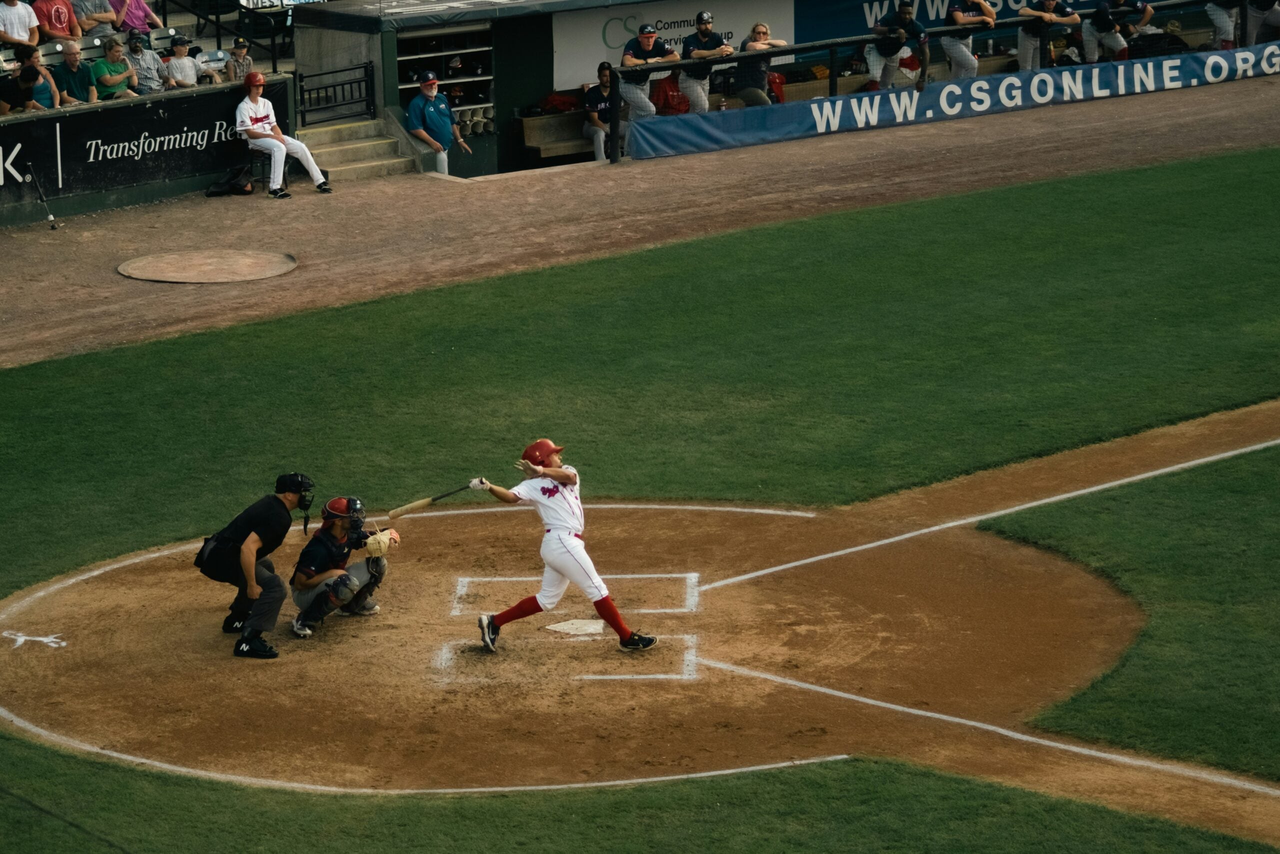 A baseball player in a red and white uniform swings a bat at home plate, while the catcher and umpire observe. Spectators and team members watch from the dugout and stands.