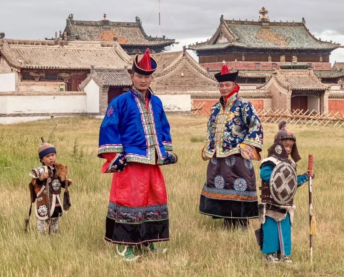 Four people in traditional Mongolian attire stand on grass, with historic temple buildings in the background. Two adults wear ornate blue robes and hats; two children wear armor and traditional clothing, holding props.