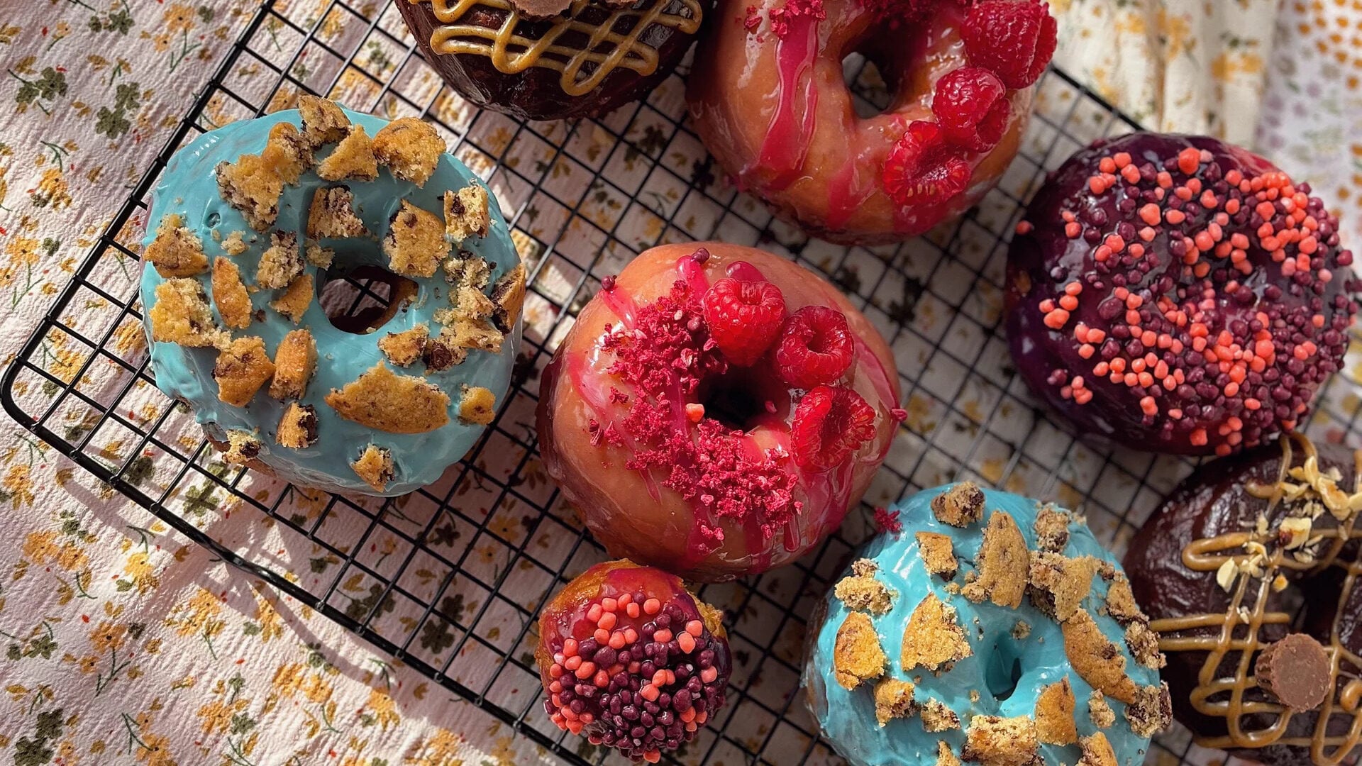 Colorful donuts topped with frosting, crumbled cookies, raspberries, and pink crunchy pearls rest on a cooling rack over a floral cloth in sunlight.
