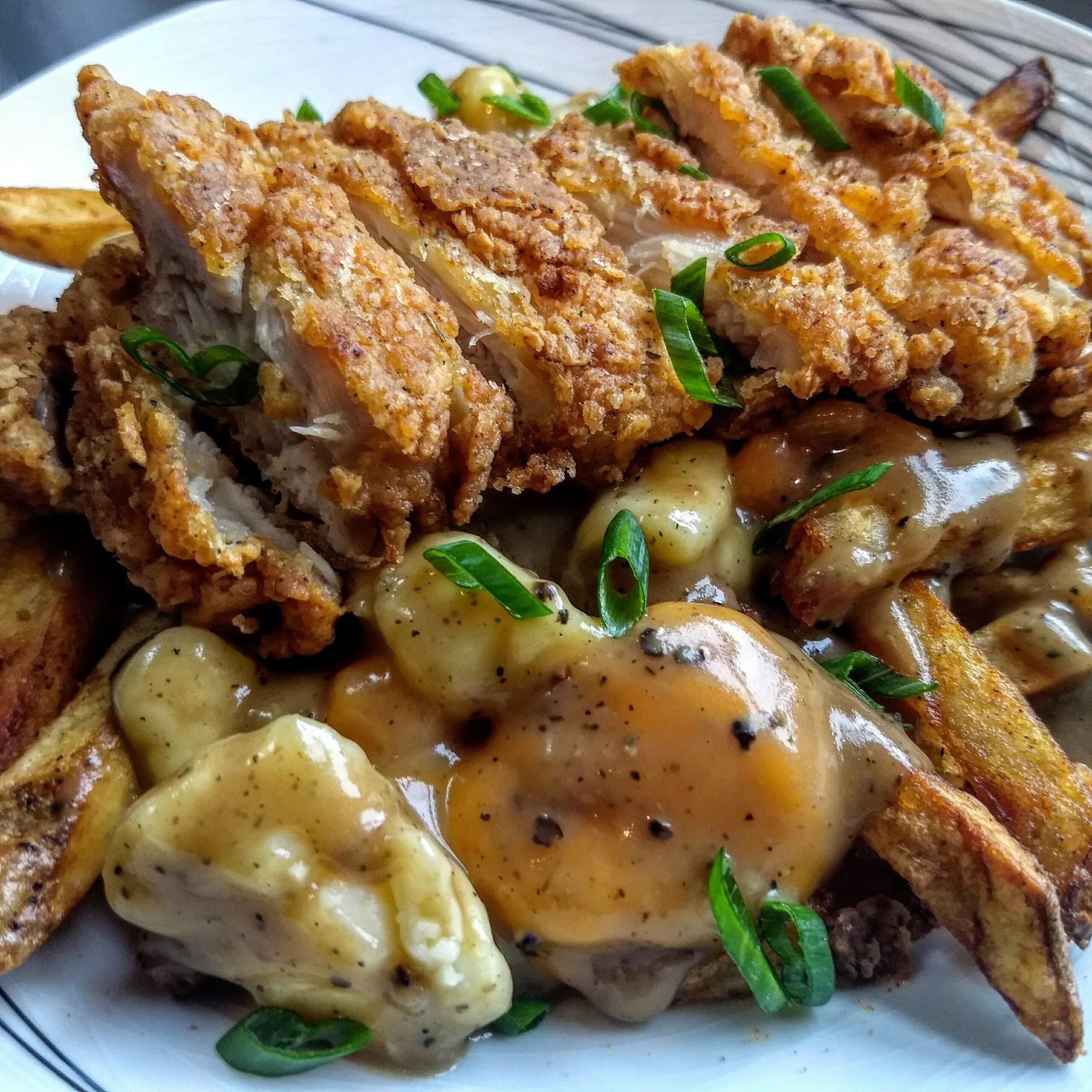 A plate of poutine topped with golden, crispy fried chicken slices, fresh green onions, cheese curds, and rich brown gravy over thick-cut fries.