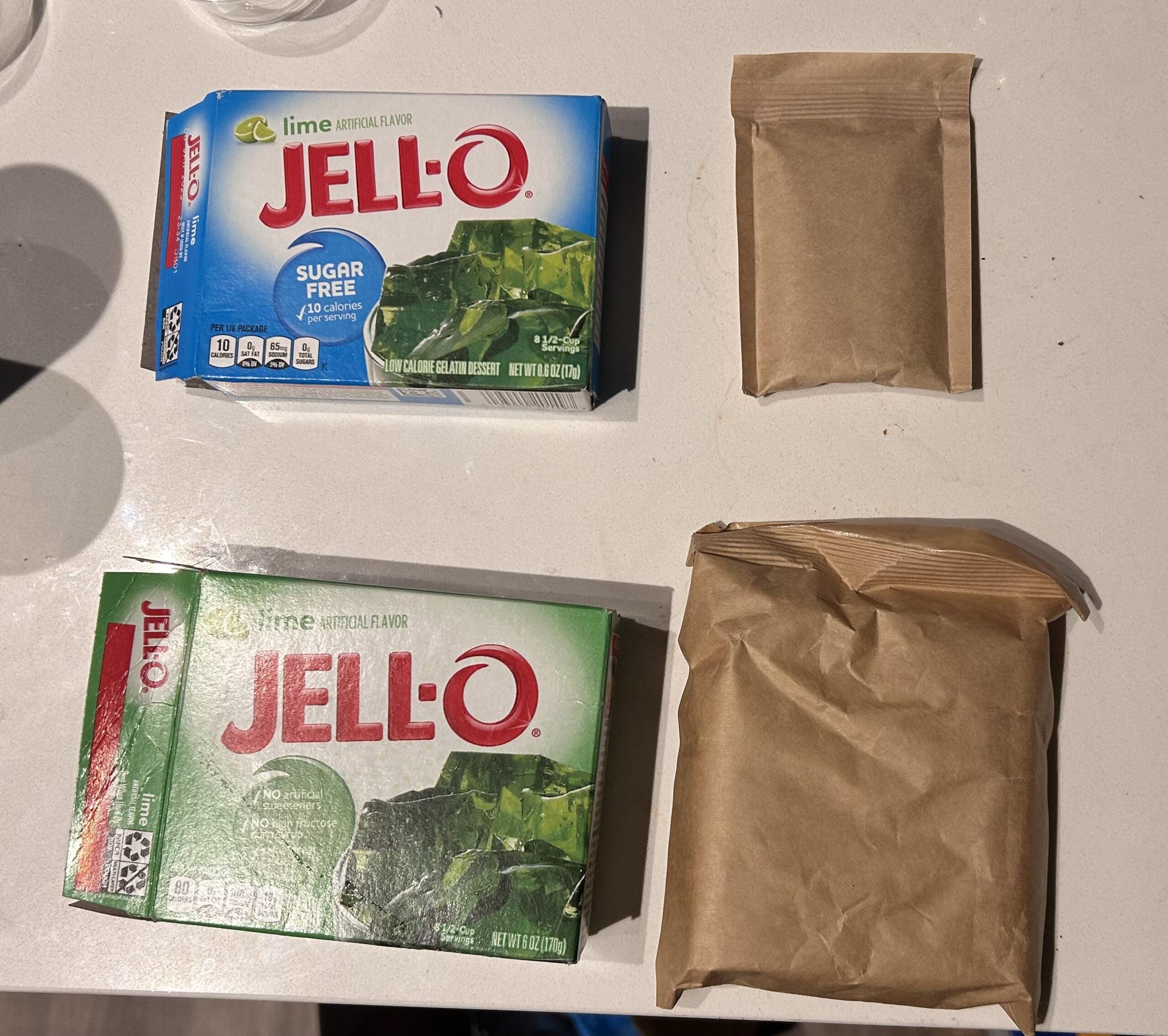 Two boxes of Jell-O lime gelatin, one sugar free and one regular, are on a white surface. Each box is next to a brown paper packet, possibly containing the gelatin mix.