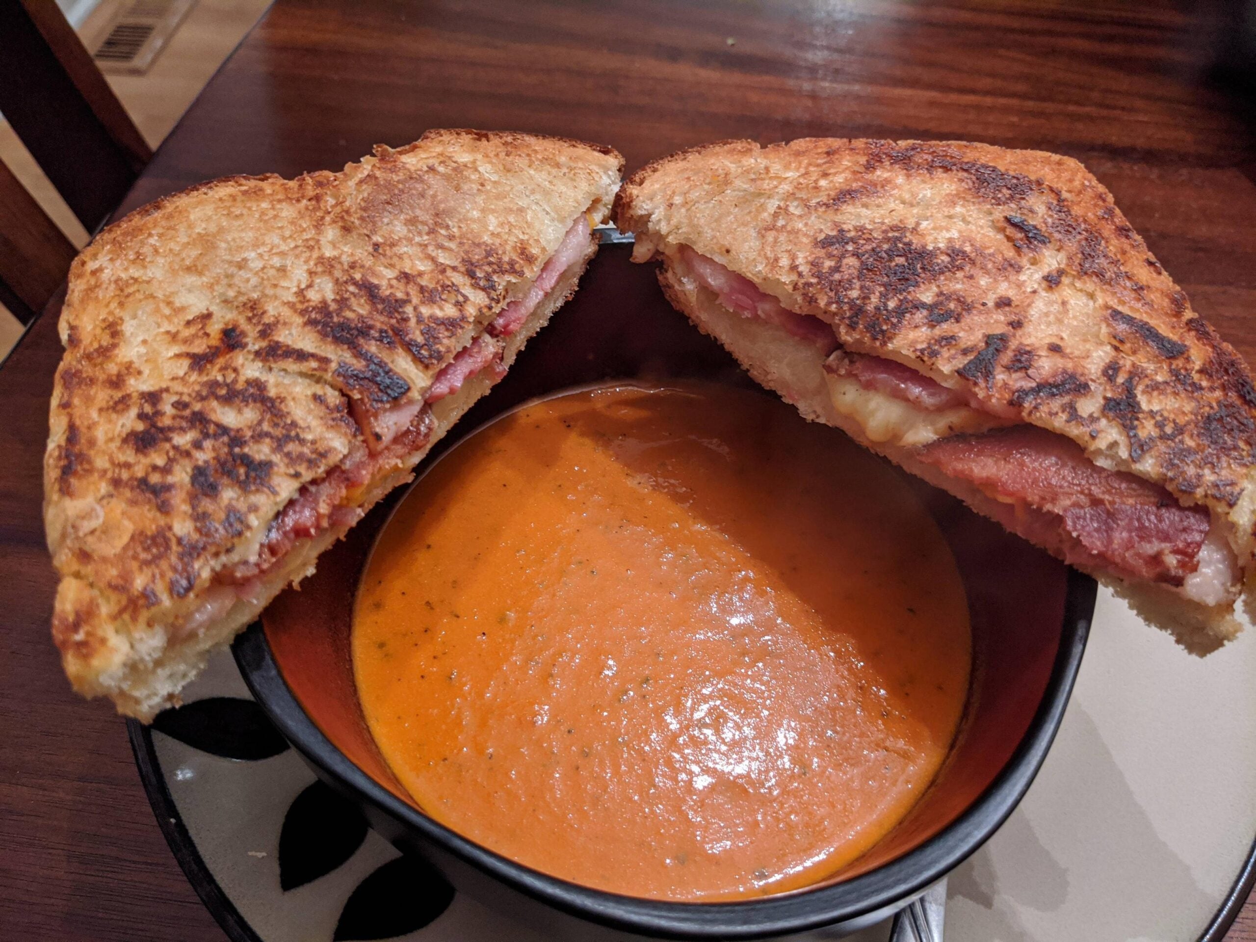 A bowl of tomato soup with a grilled cheese sandwich, cut in half and resting on the bowl’s rim. The sandwich appears to contain bacon and melted cheese. The meal is set on a dark wooden table.