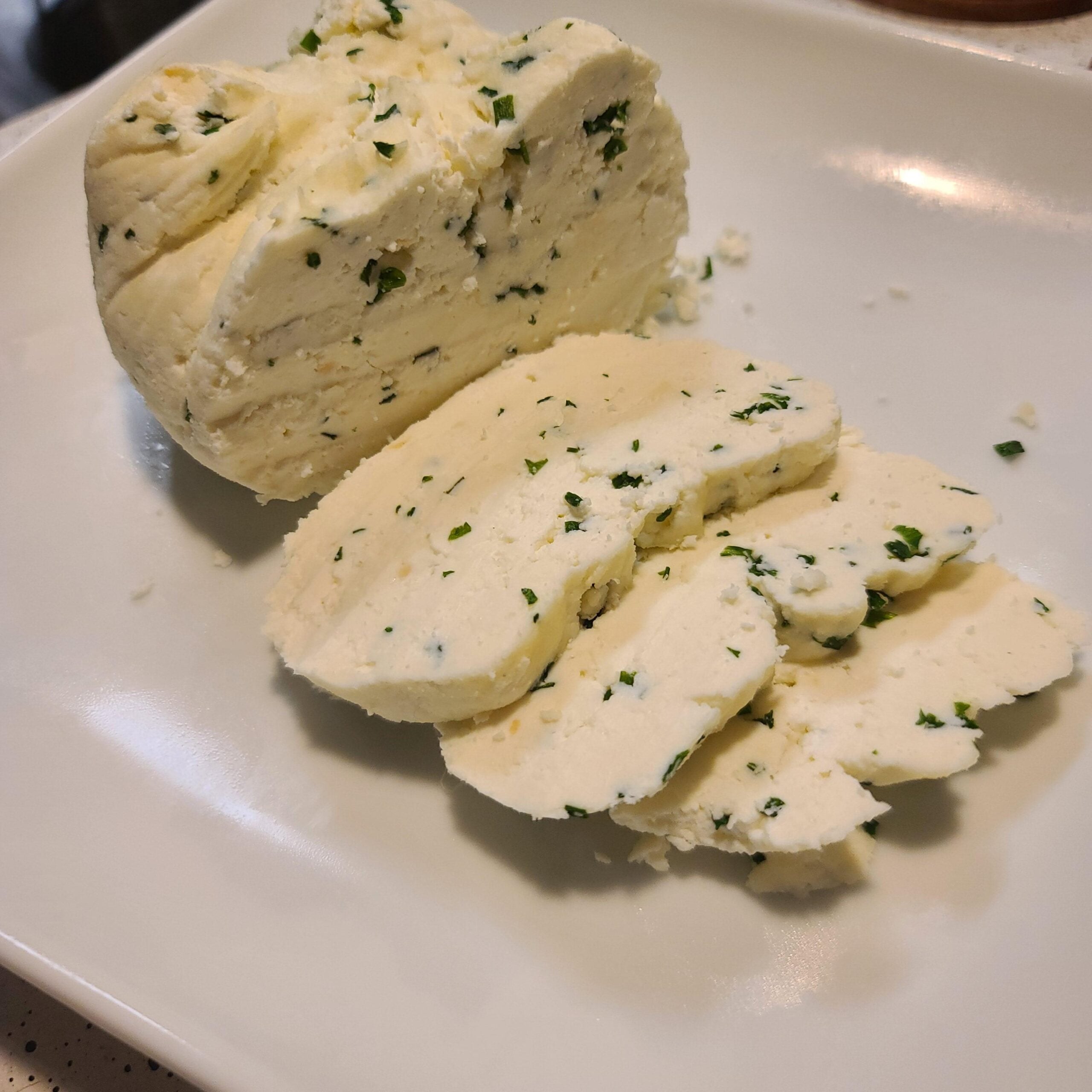 A block of white cheese with herbs is partially sliced on a white plate. The cheese appears soft and crumbly, with green herb pieces distributed throughout.