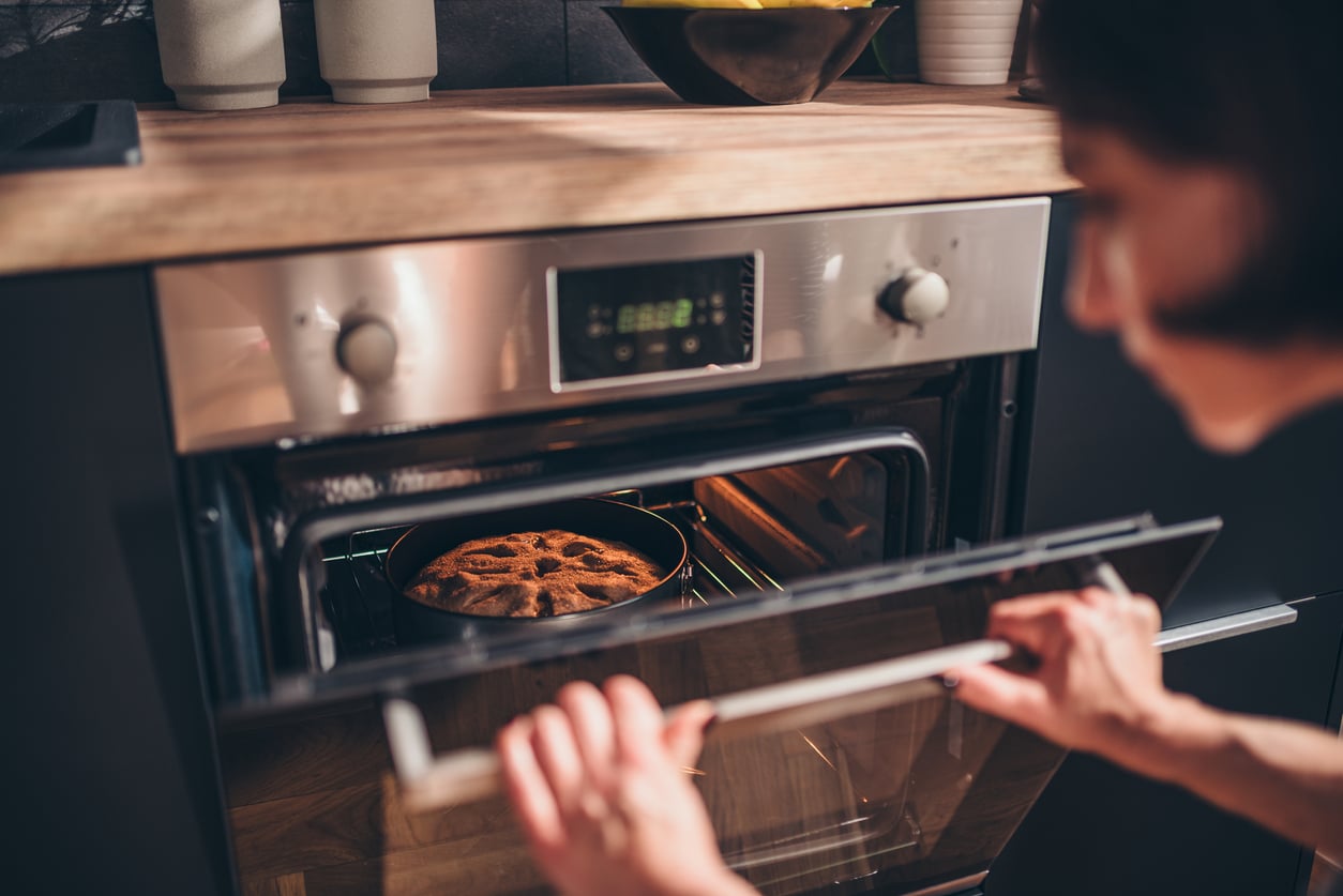 A person opens an oven door to check on a cake baking in a round pan. The kitchen counter above holds a bowl of fruit and other kitchen items.