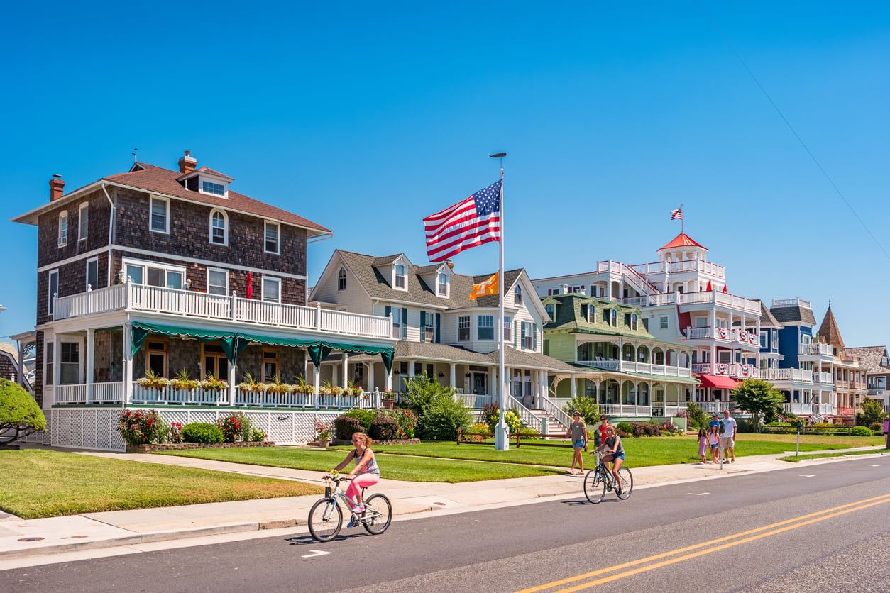 People ride bicycles on a sunny street lined with colorful, historic houses and an American flag on a pole, evoking the charm of nostalgic travel destinations. The sky is clear and blue.