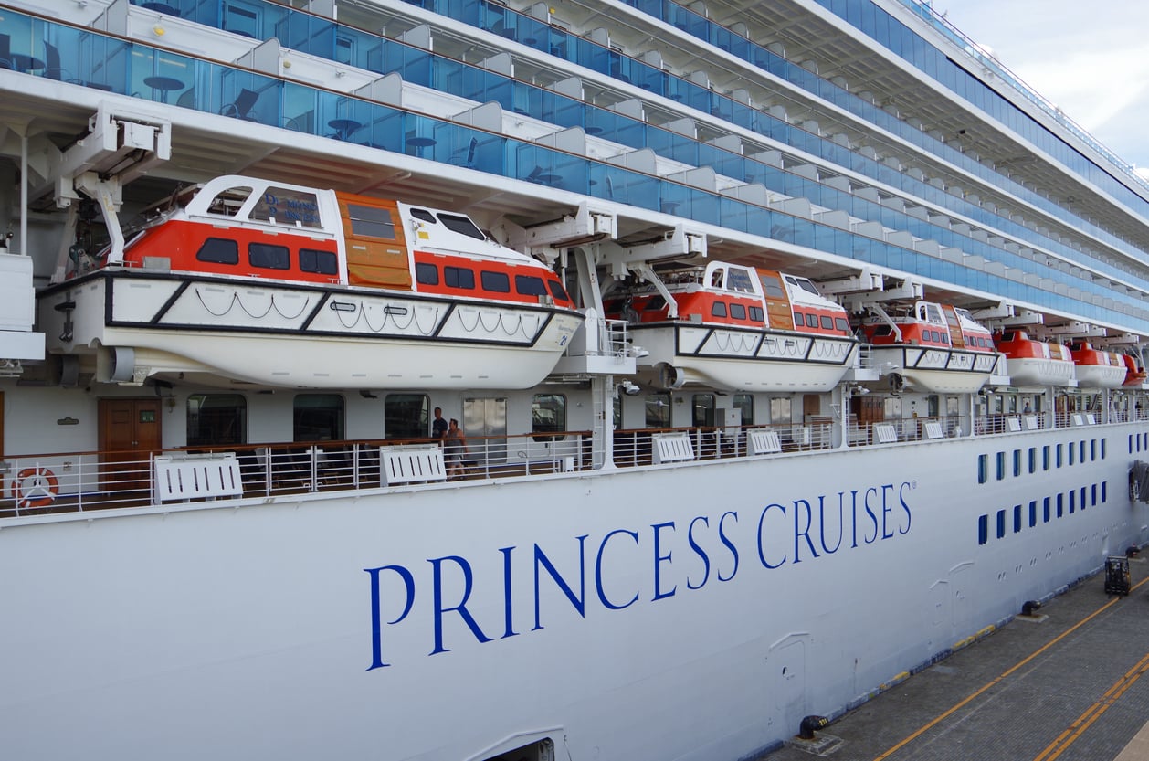The side of a white Princess Cruises ship docked at a port, showing several orange and white lifeboats secured above the deck and large windows above them.