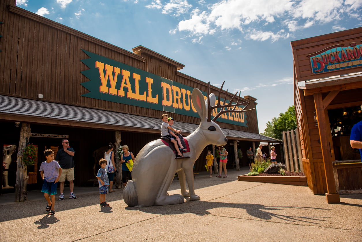People walk outside Wall Drug. A boy sits on a giant jackalope statue while others look on. The weather is sunny with a few clouds, and the scene is lively with visitors.