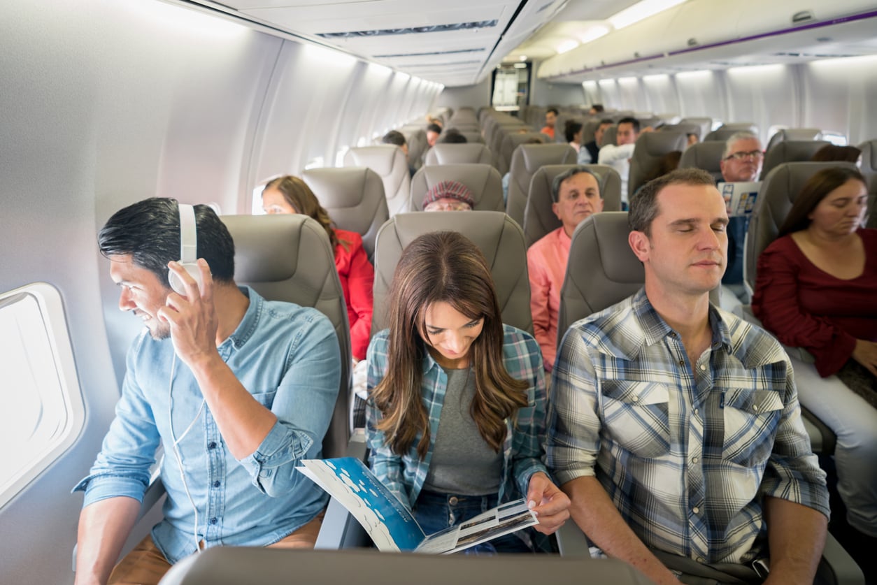 Passengers sit in airplane seats; one man looks out the window with headphones, a woman reads a magazine, and another man sits with his eyes closed. Other travelers are visible in the background.