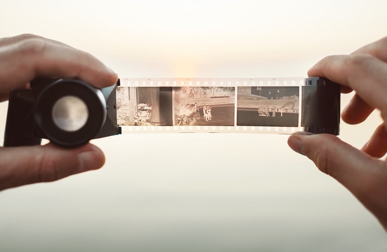 Close-up of two hands holding a strip of photographic film negatives against a bright, blurred background, with a magnifier held against the film to view the images in detail.