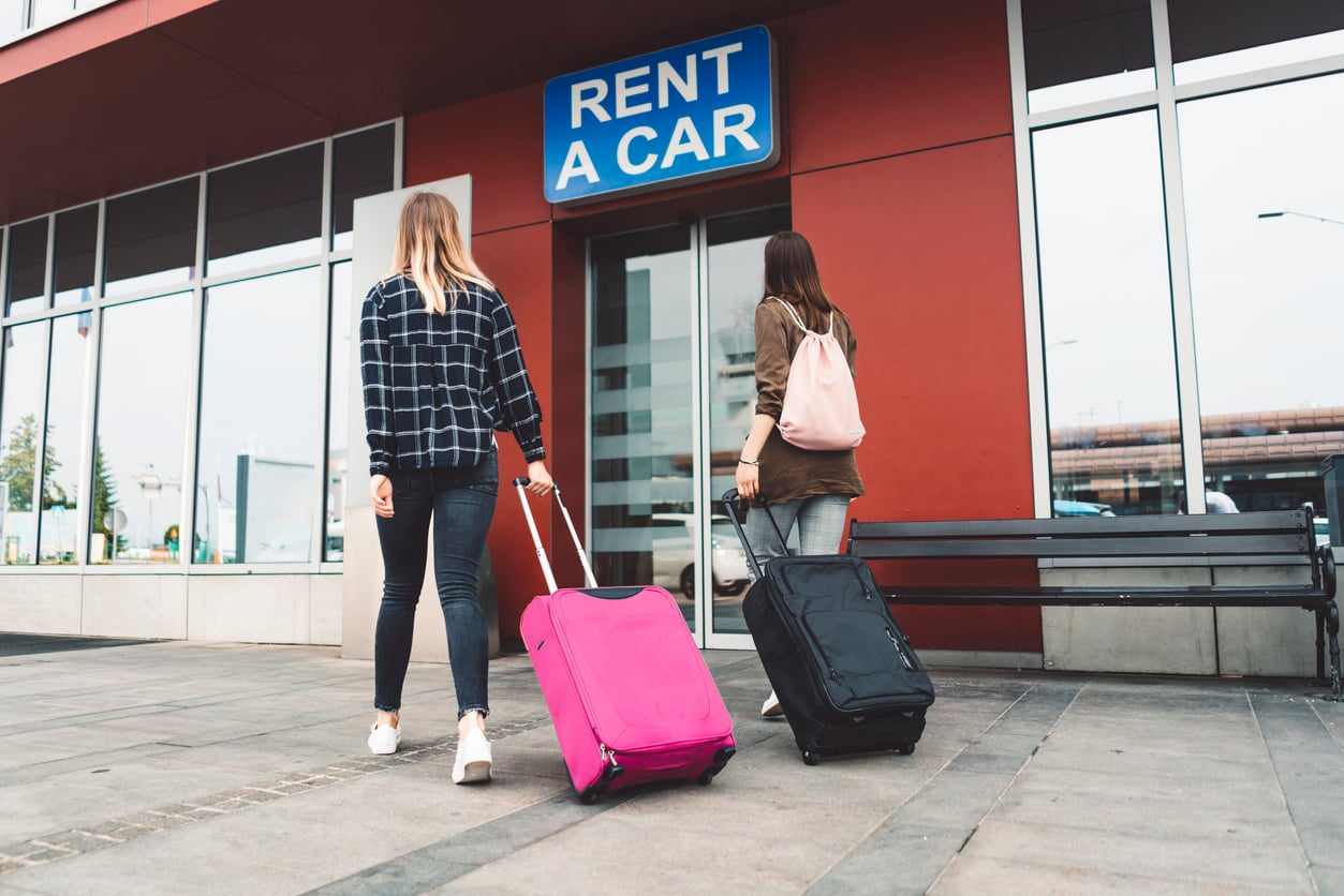 Two women with rolling suitcases walk toward a “Rent a Car” office, located in a modern building with large windows. One suitcase is bright pink and the other is black.