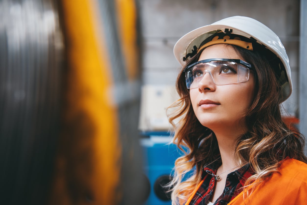 A woman wearing a white hard hat, safety glasses, and an orange vest looks up thoughtfully in an industrial setting, with blurred machinery in the background.
