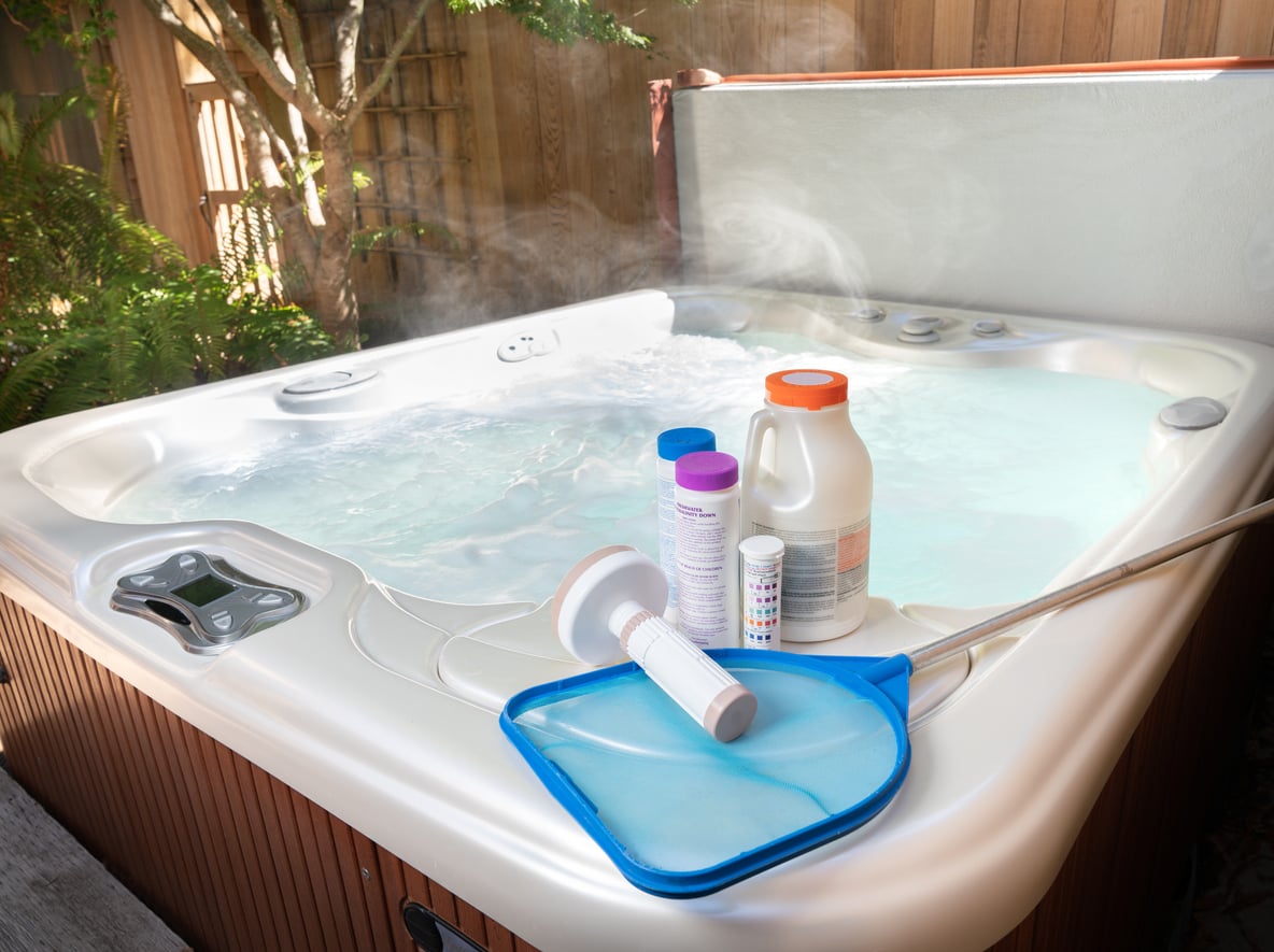 A steaming hot tub outdoors with a skimmer net, floating dispenser, chlorine tablets, test strips, and other cleaning supplies placed on its edge.
