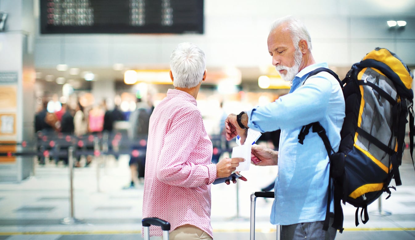 Two older adults with luggage stand in an airport terminal, demonstrating their travel habits. One checks the time on his watch while the other looks at the departure board. The scene is busy, with people and signs blurred in the background.