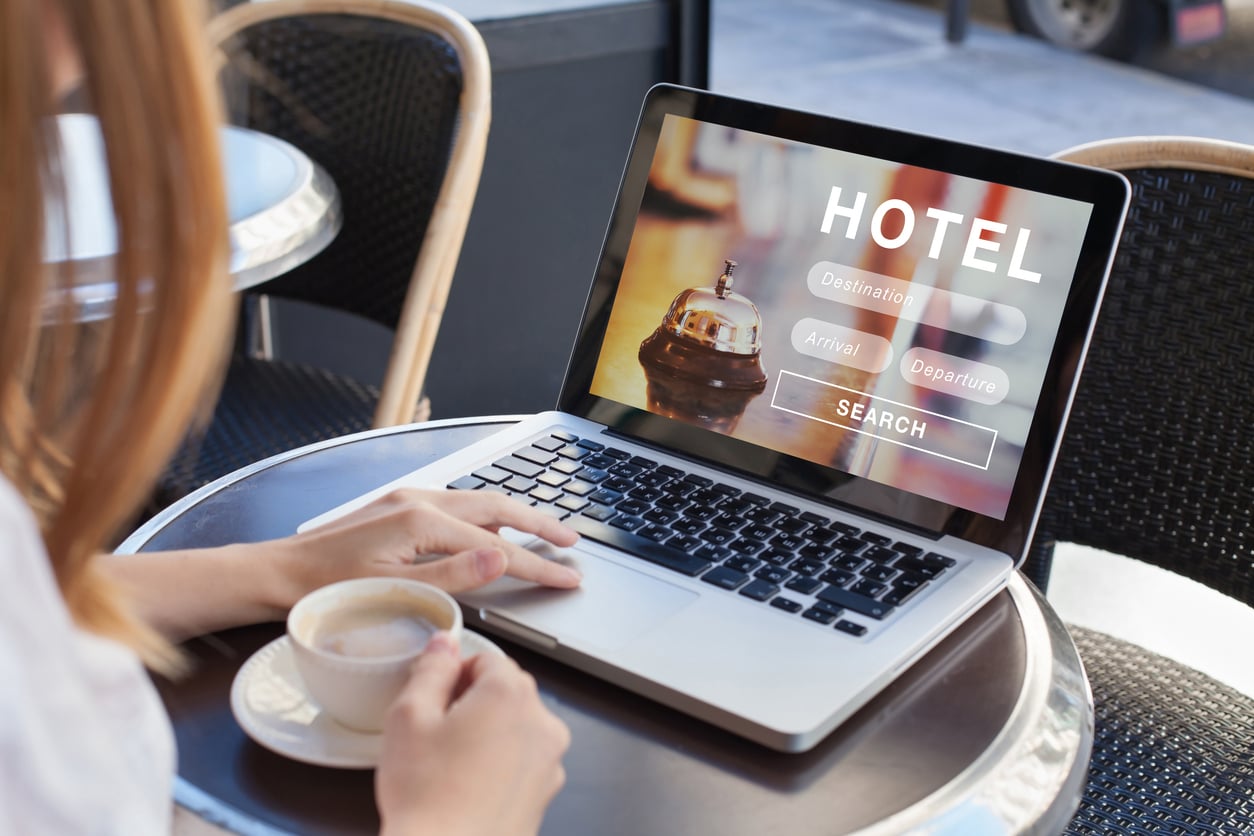 A person with a cup of coffee is using a laptop at a café. The laptop screen displays a hotel booking website with search options for destination, arrival, and departure dates.