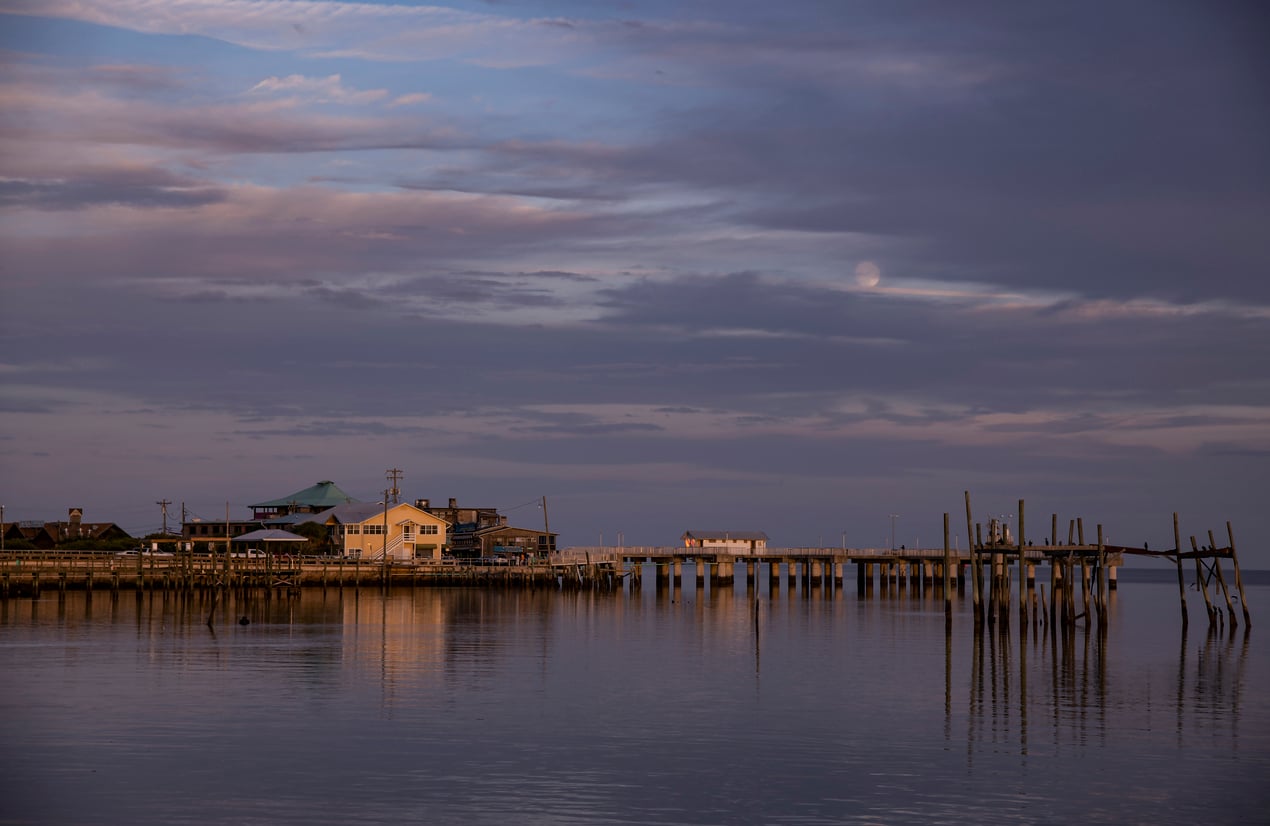 A pier extends over calm water at sunset, with houses and posts reflecting on the surface. Clouds and a faint moon are visible in the sky.