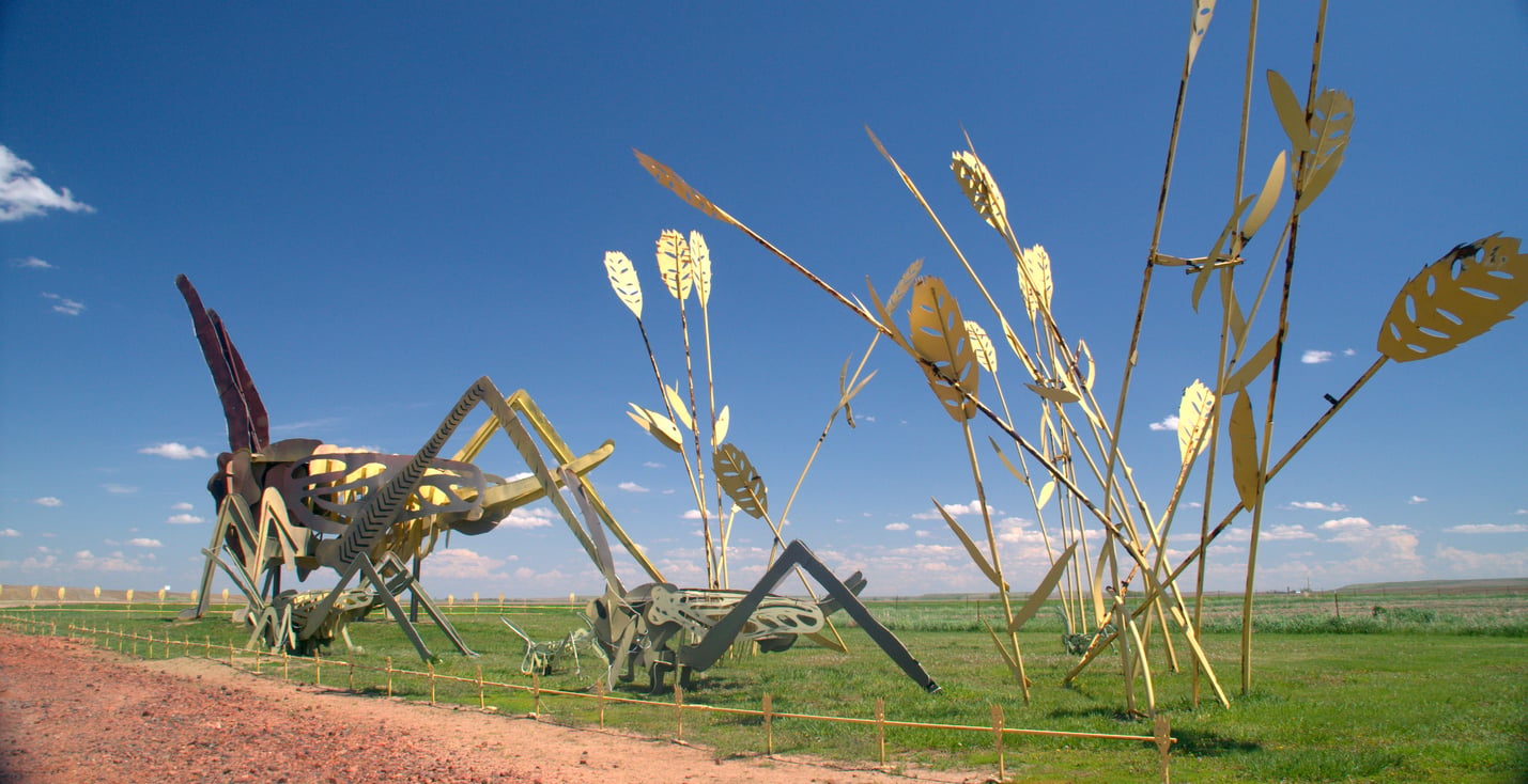 Large metal sculptures of grasshoppers and tall wheat stand in a grassy field under a bright blue sky, with a gravel path nearby.