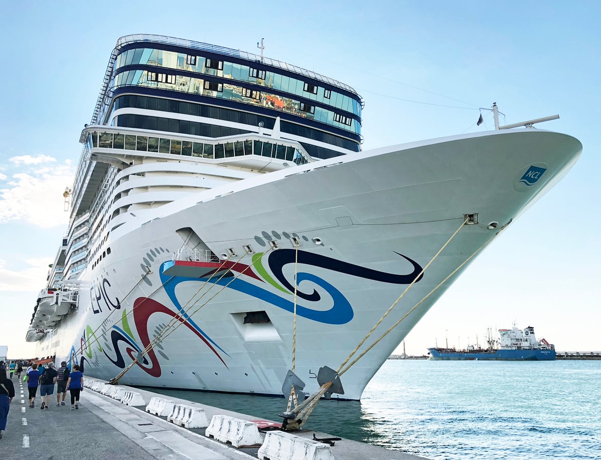 A large white cruise ship with colorful designs is docked at a port. People walk nearby on the pier, and smaller ships are visible in the background on the water.