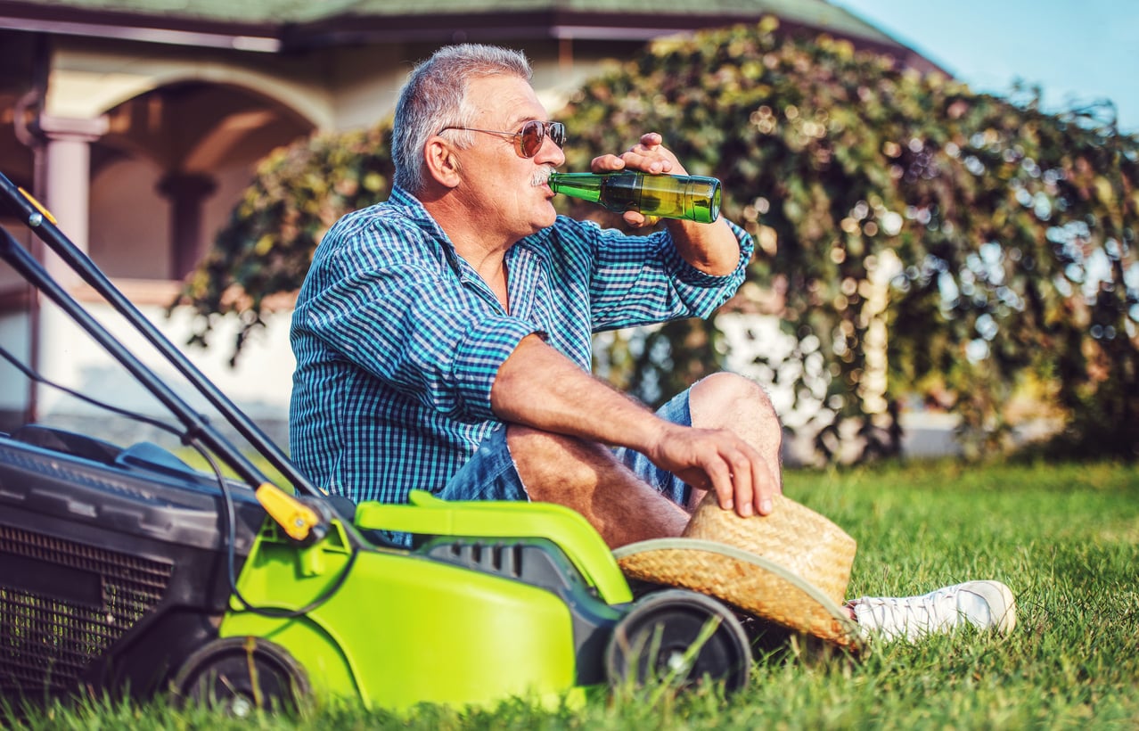 An older man sits on grass beside a green lawnmower, holding a straw hat and drinking from a green bottle. He wears sunglasses and a plaid shirt, relaxing in front of a house on a sunny day.