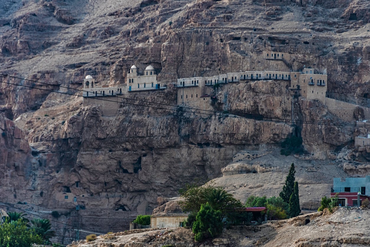 A long, light stone monastery with domes is built into a rugged, rocky cliff, surrounded by arid hills, sparse vegetation, and a few trees and buildings at the base. Power lines cross in front.