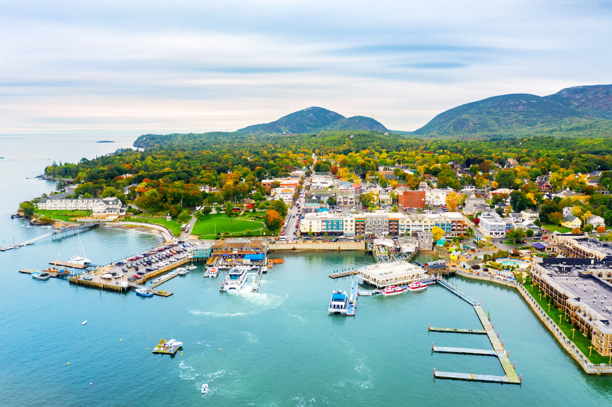 Aerial view of a coastal town with a marina, colorful buildings, green trees, and mountains in the background under a cloudy sky—a perfect destination to explore after age 50.