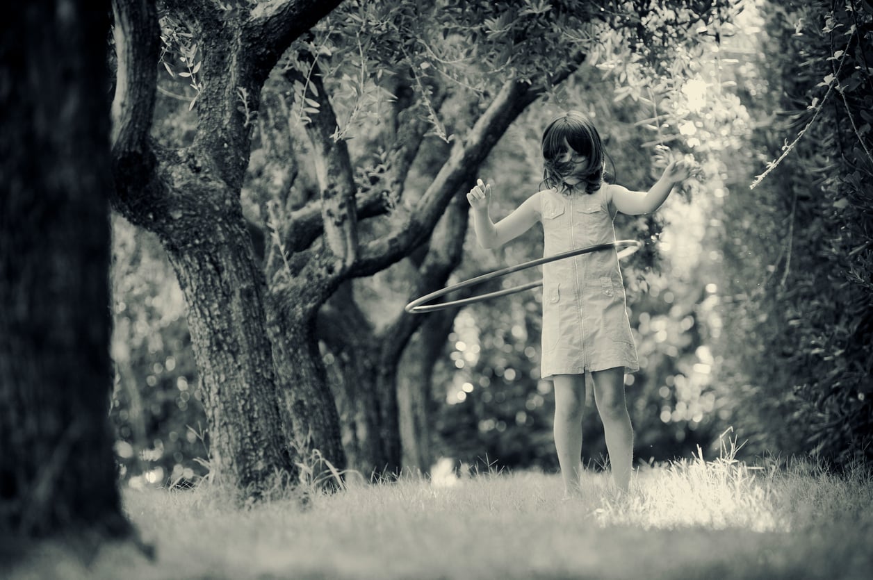 A young girl in a dress plays with a hula hoop among trees in a sunlit, grassy area. The image is in black and white, creating a nostalgic, peaceful atmosphere.