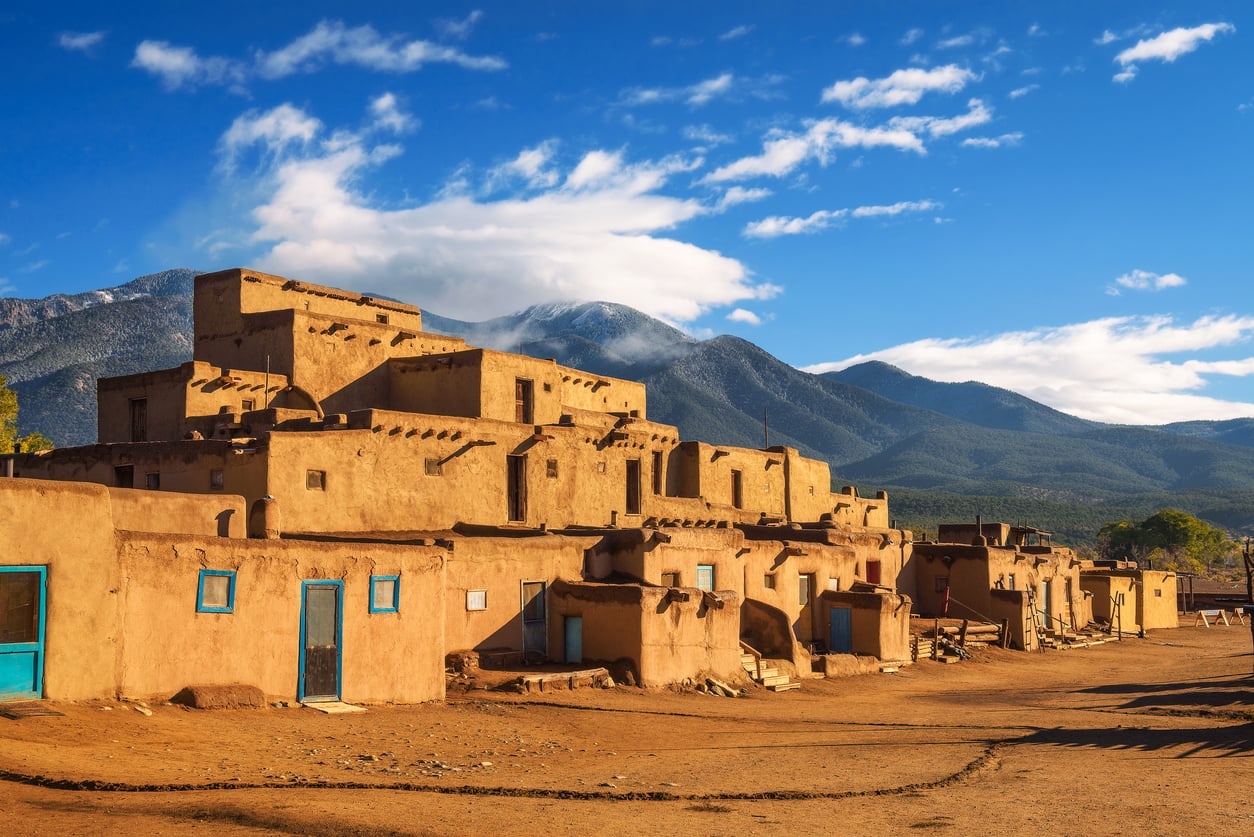 Adobe-style pueblo buildings with blue doors sit in front of tree-covered mountains under a bright blue sky with scattered clouds, evoking the charm and beauty found in some of the best fall towns in America.
