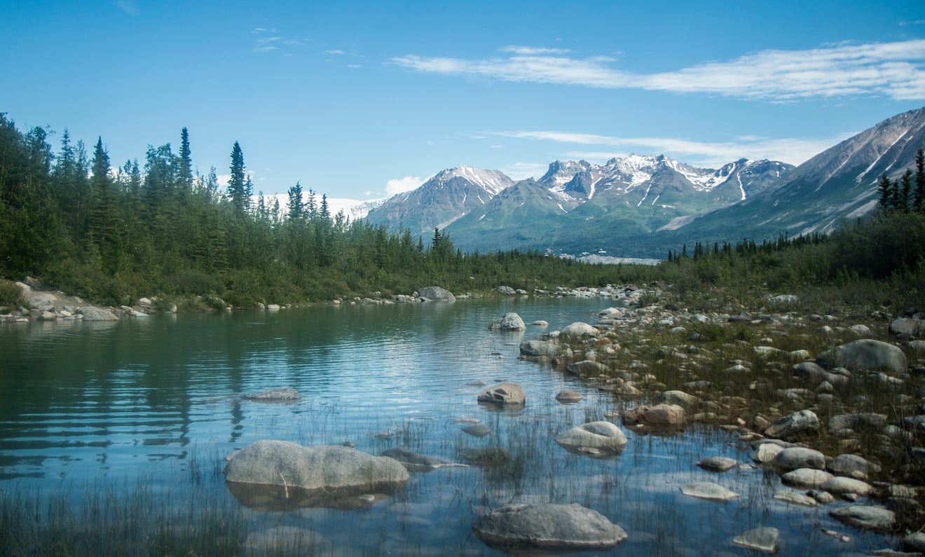 A clear, shallow river flows through a forested valley with rocks and trees, leading toward distant snow-capped mountains under a blue sky with scattered clouds—a scene reminiscent of the breathtaking landscapes featured in many travel facts about the United States.